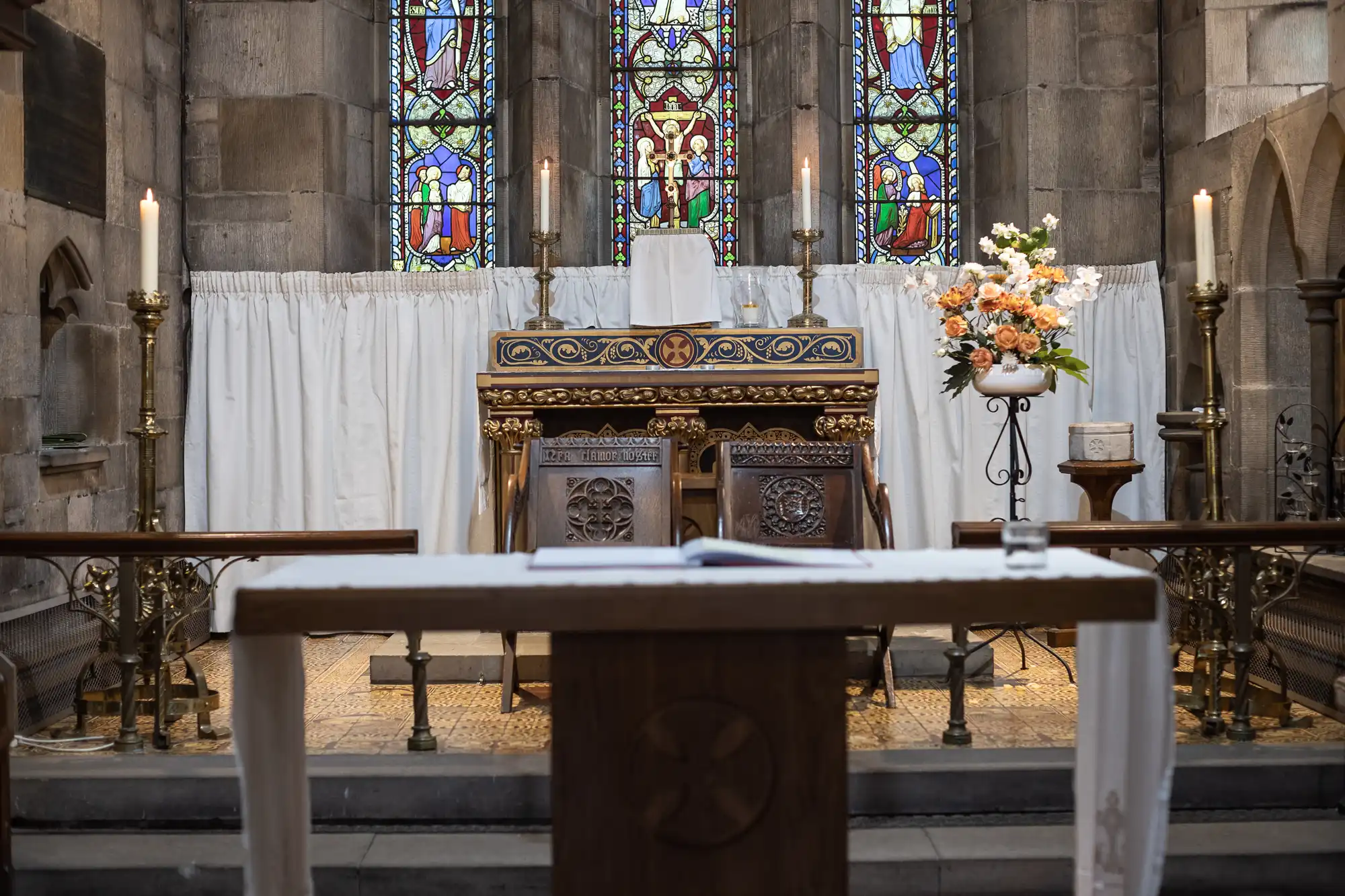 A church altar with stained glass windows, ornate wooden furniture, lit candles, and floral arrangements.