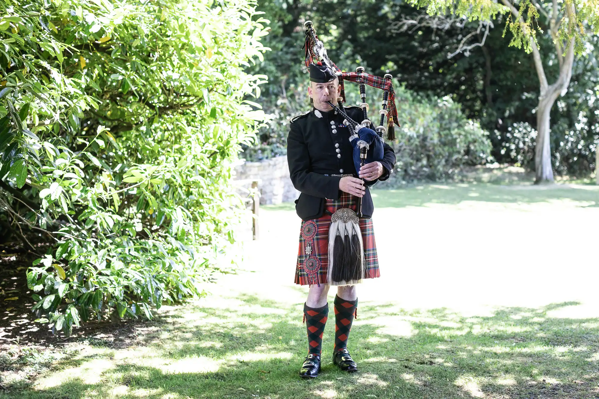 A person in traditional Scottish attire plays the bagpipes outdoors on a grassy area with trees and sunlight in the background.