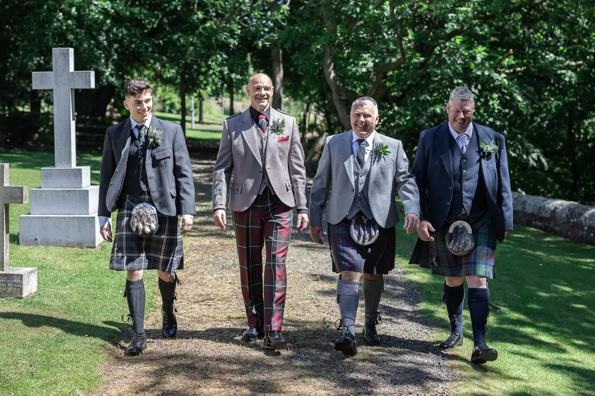 Four men in kilts and jackets walk on a path in a cemetery, smiling and talking.