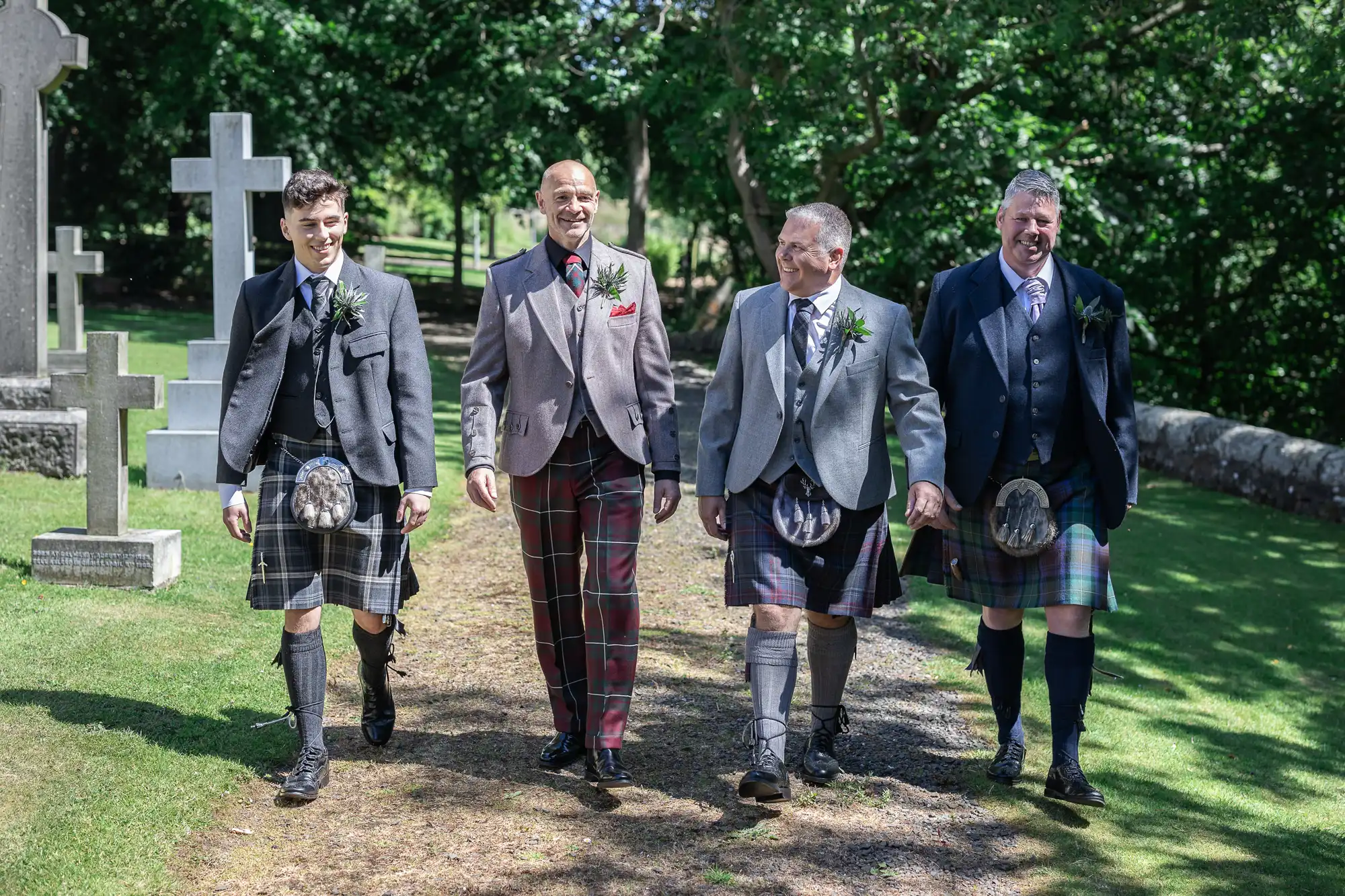 Four men dressed in kilts and jackets walk together outdoors on a sunny day, surrounded by greenery and some gravestones.