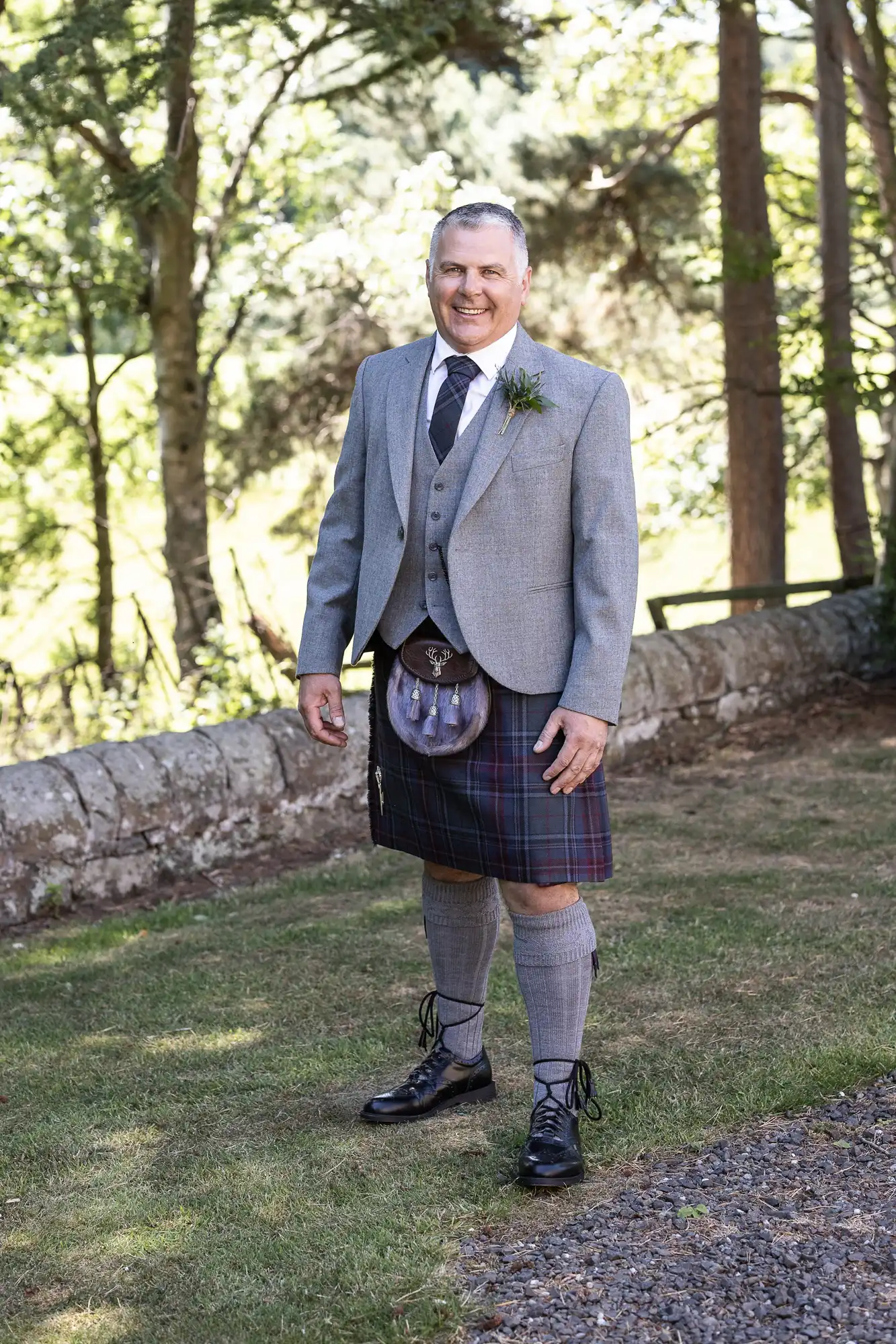 A man stands in a garden wearing a grey jacket, black vest, tie, and a tartan kilt with matching socks and black shoes, posing for the camera. Trees and a stone wall are in the background.