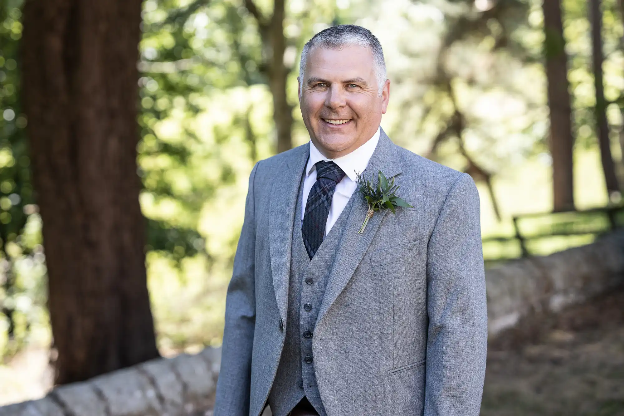 A man wearing a gray suit with a boutonniere stands outdoors in front of a forested background, smiling at the camera.