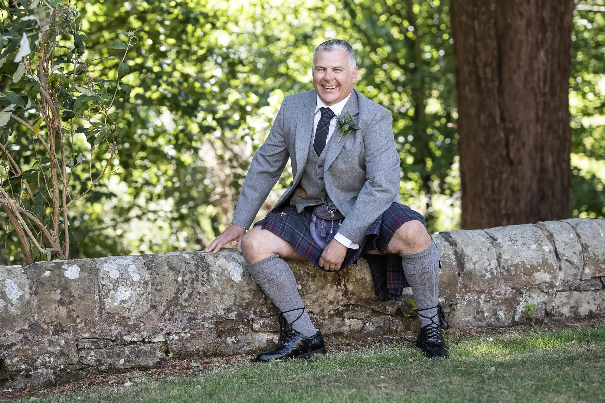 A man wearing a grey jacket, tie, and kilt sits on a stone wall in an outdoor, wooded area.