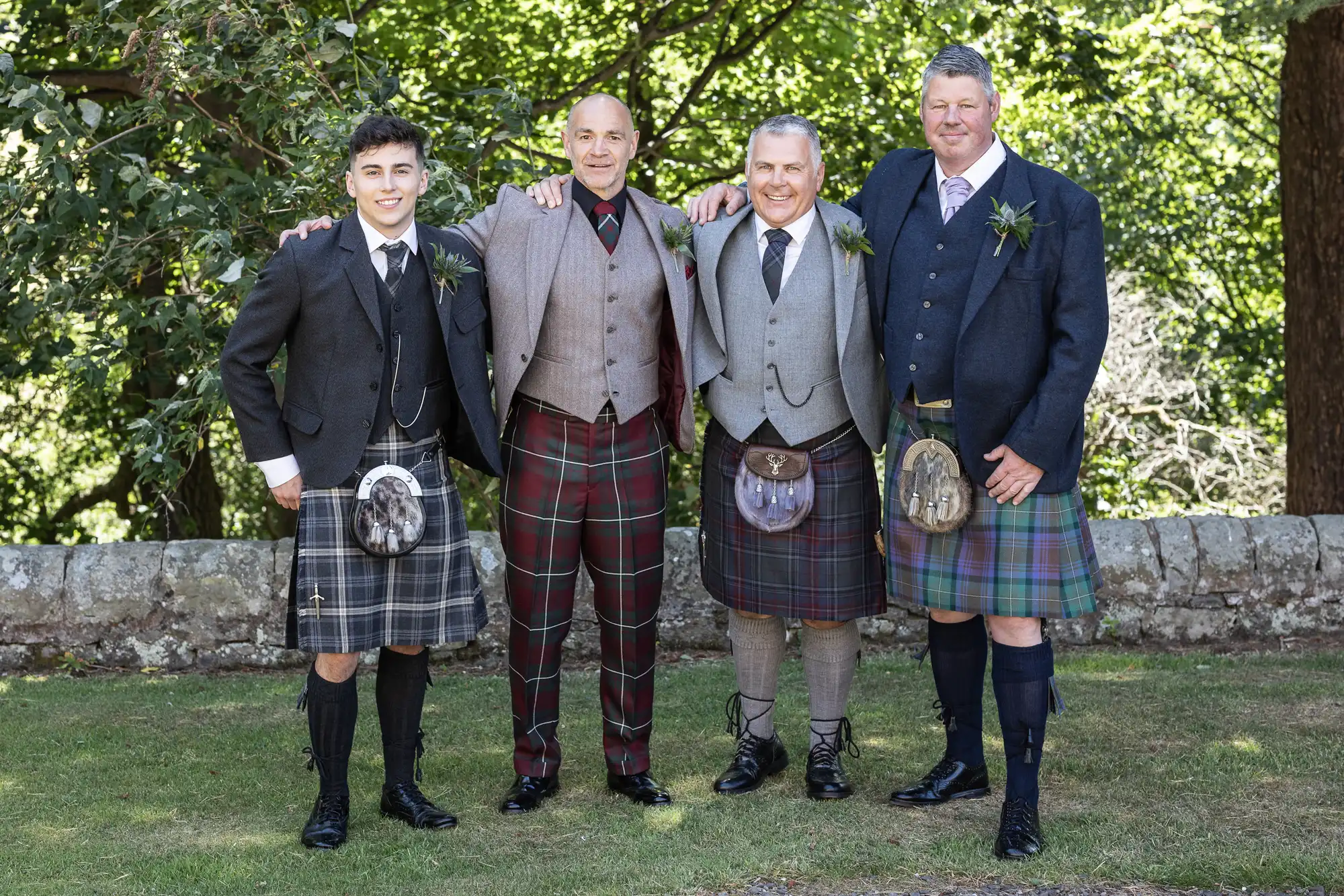 Four men in kilts and formal jackets stand side by side outdoors, smiling at the camera. Trees and a stone wall can be seen in the background.