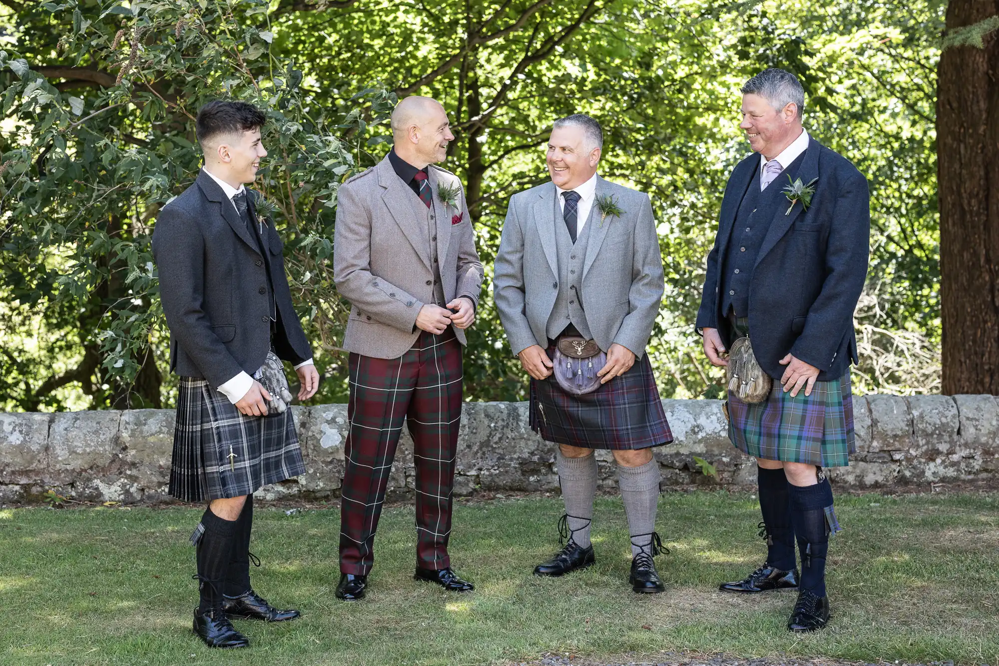 Four men in kilts are standing outdoors, conversing and smiling. They are wearing traditional Scottish attire with jackets and sporrans. Trees and a stone wall are visible in the background.