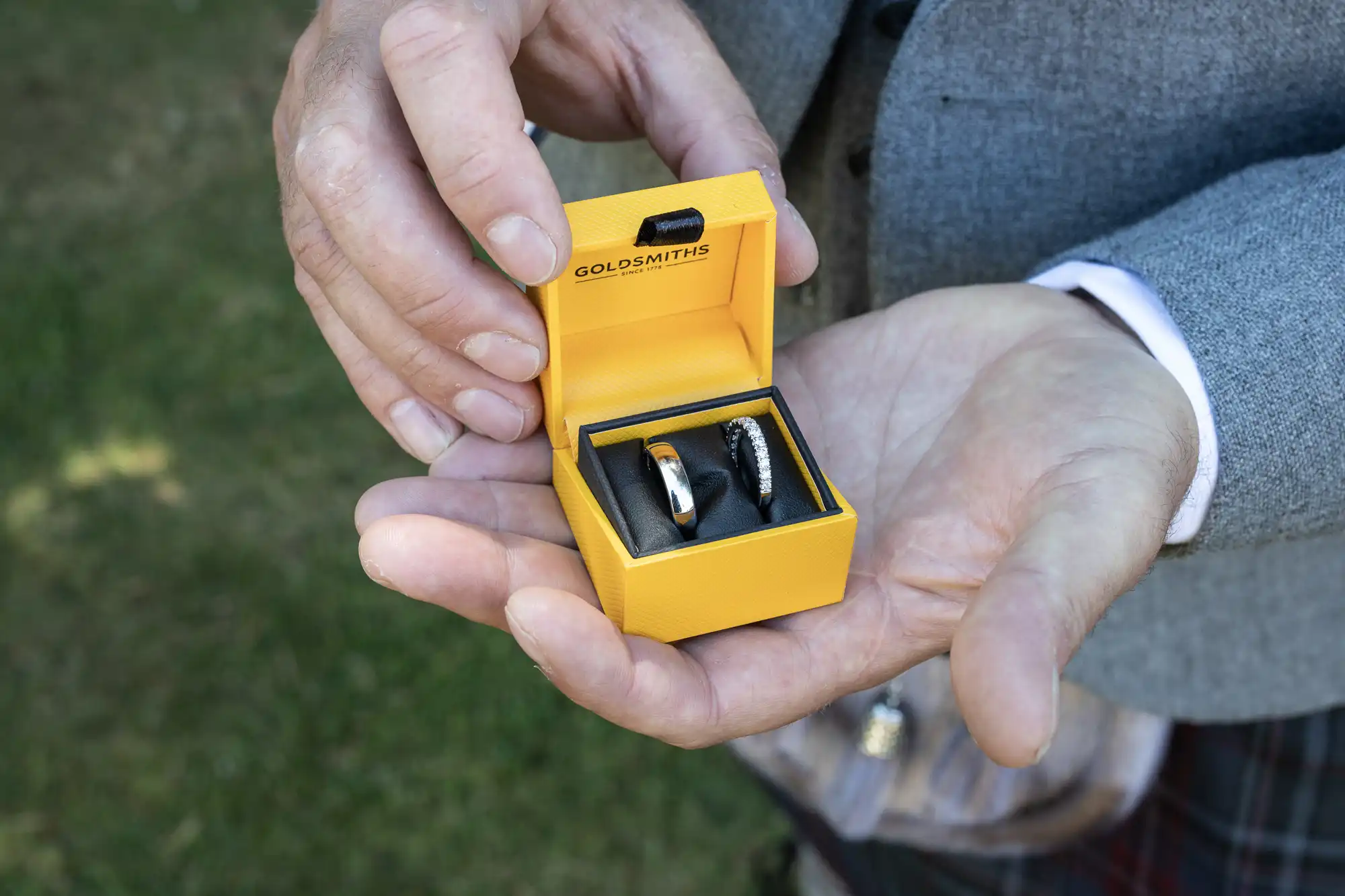 A person in a grey jacket holds an open yellow Goldsmiths branded box containing two rings.