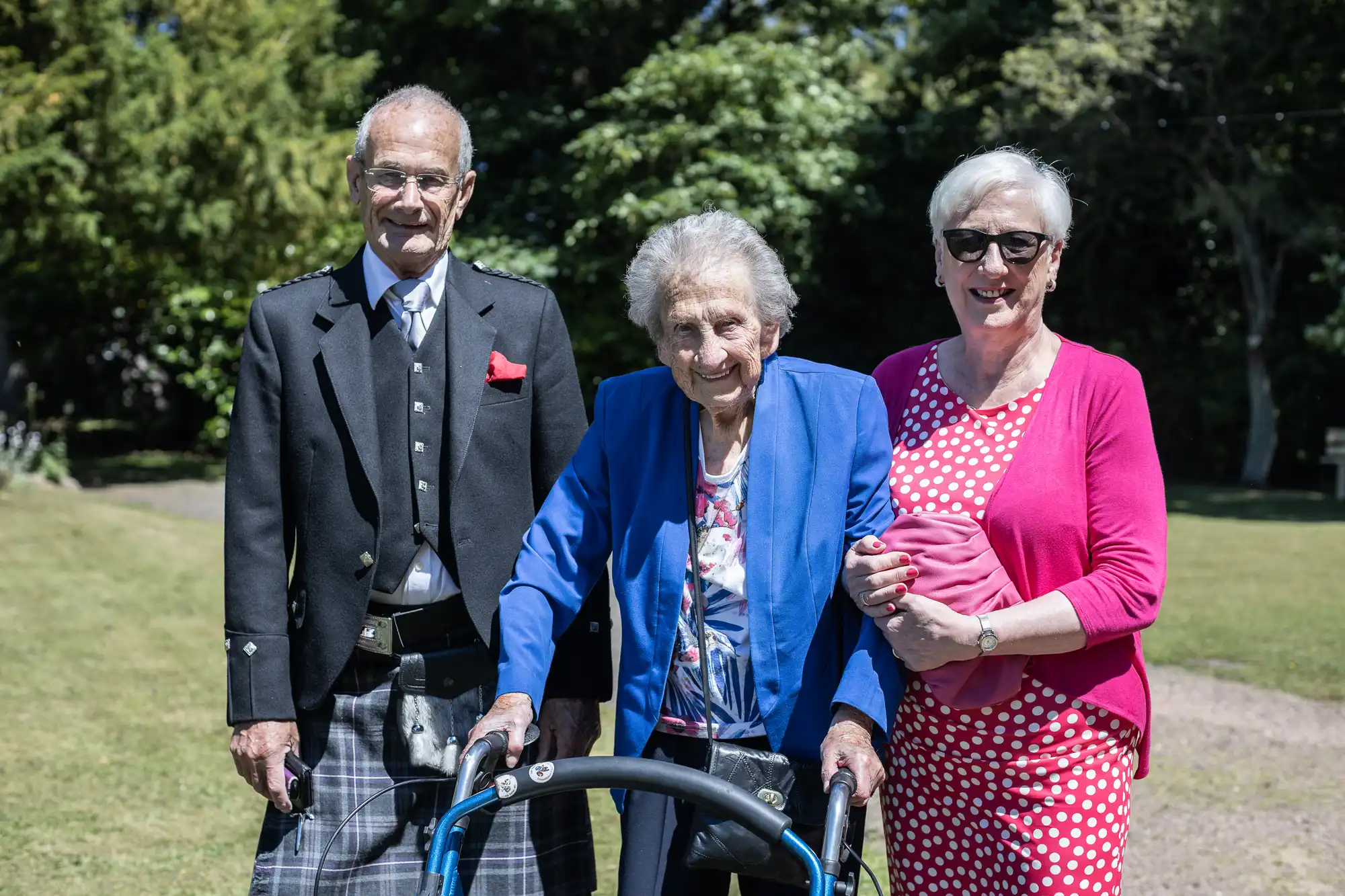 Three elderly individuals posing outdoors; a man in traditional attire, an elderly woman with a walker, and a woman in a pink cardigan and sunglasses.