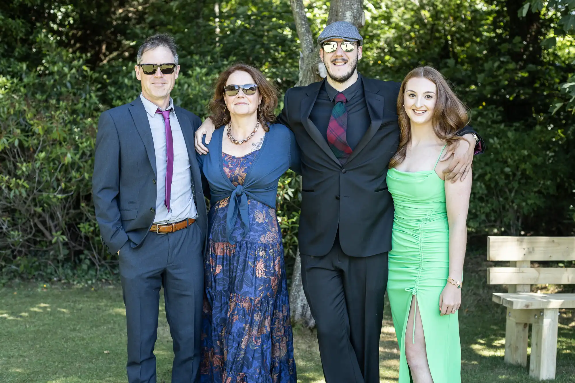 Four people posing outdoors in formal attire, standing in front of a tree. Two men and two women, all wearing sunglasses. One woman is in a green dress while the others are in suits or a patterned dress.