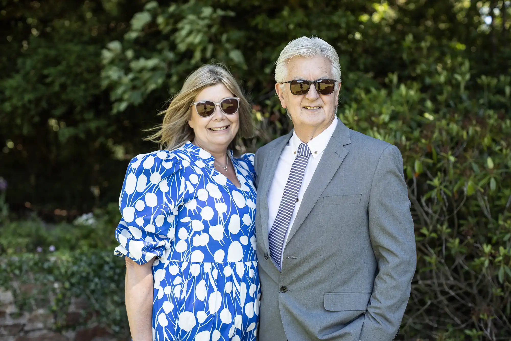 A woman in a blue and white dress and a man in a gray suit, both wearing sunglasses, stand together outside with greenery in the background.