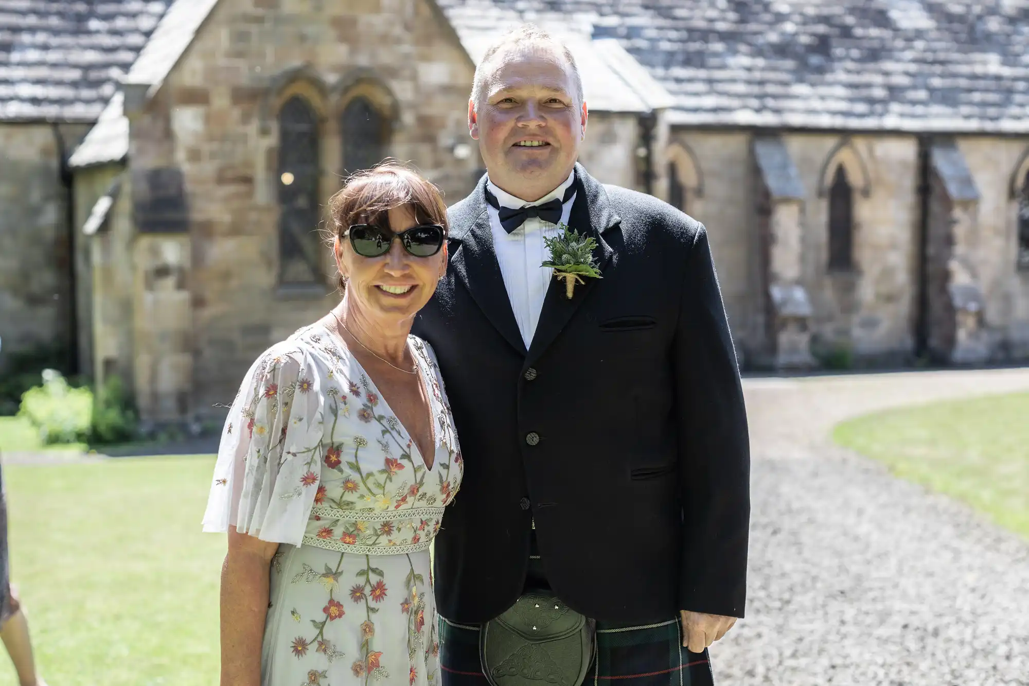 A man in formal attire and a woman in a floral dress pose together outside a stone building on a sunny day.