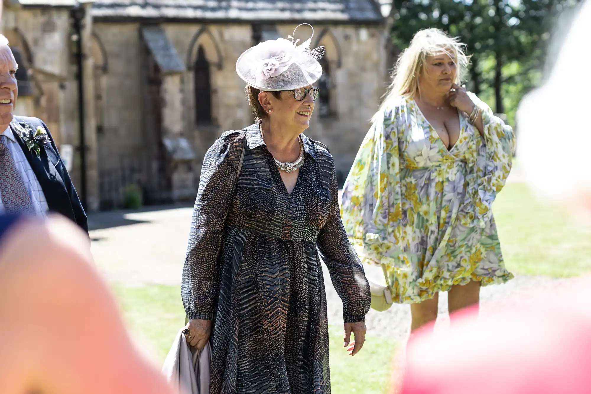 Two people dressed in formal attire stand outside a historic building on a sunny day. One person wears a patterned dress and hat, and the other wears a floral dress.