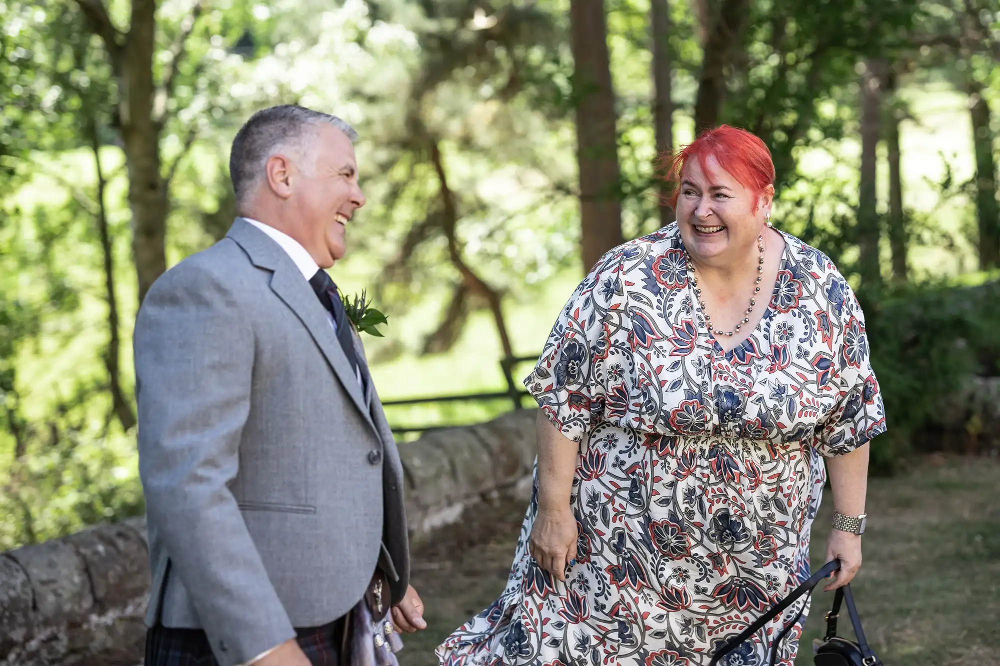 A man in a grey suit and woman in a patterned dress with red hair laugh together in an outdoor setting with trees in the background.