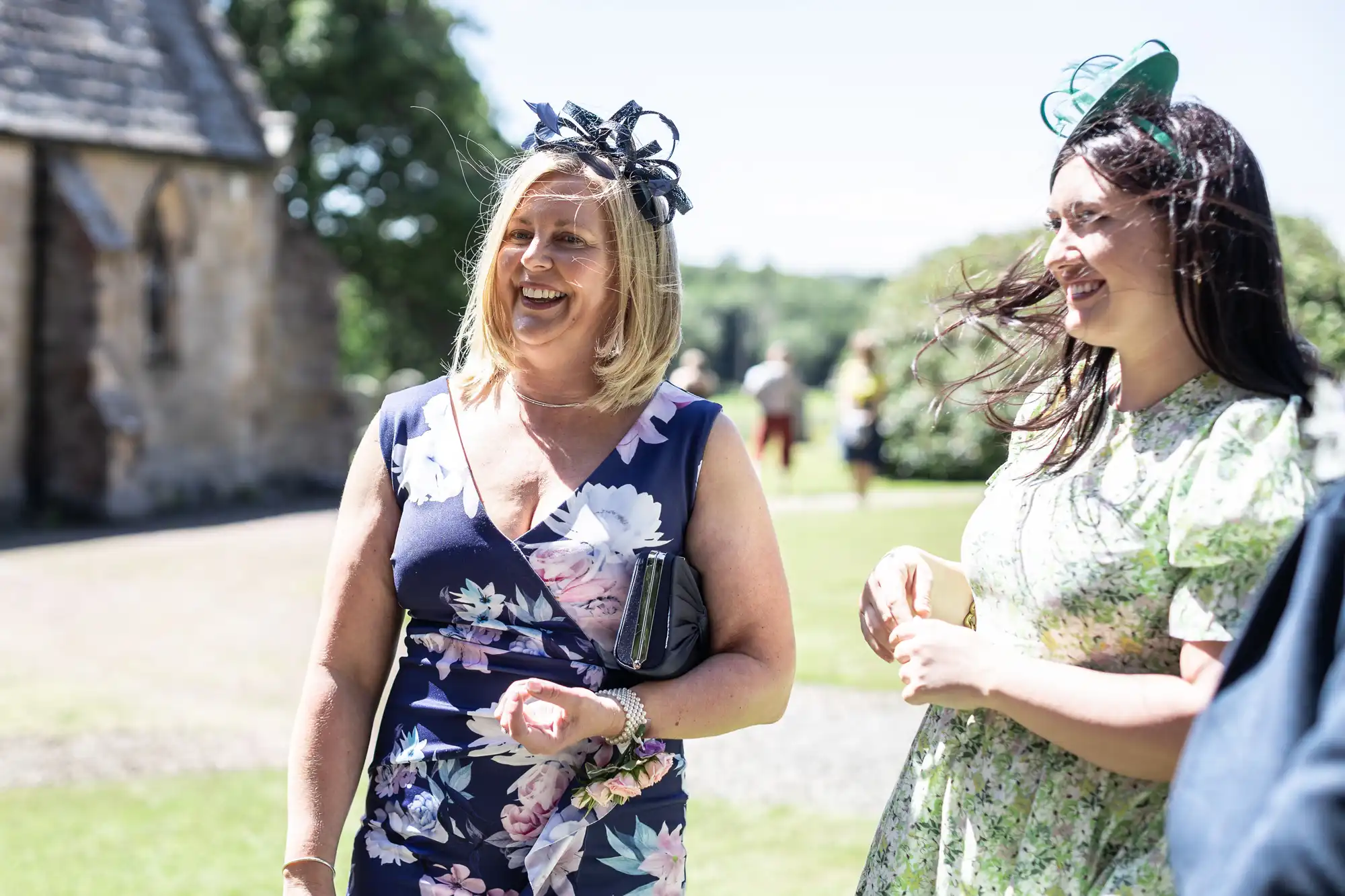 Two women wearing floral dresses and fascinators smile and converse outdoors on a sunny day. The background includes a stone building and other people.