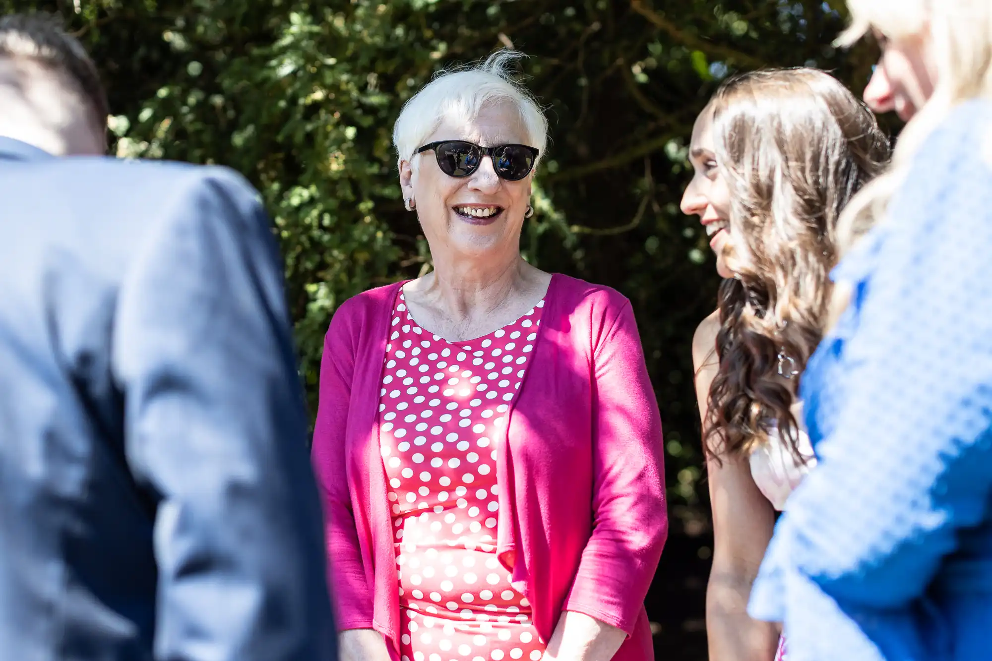 An elderly woman wearing sunglasses and a pink polka dot dress stands outside, smiling and chatting with three other people.