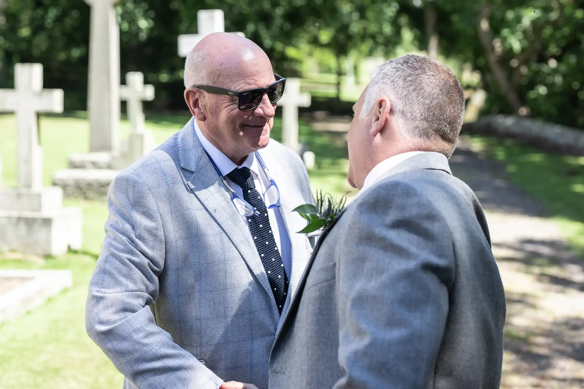 Two men in suits shake hands and smile in a sunny outdoor setting with gravestones visible in the background.