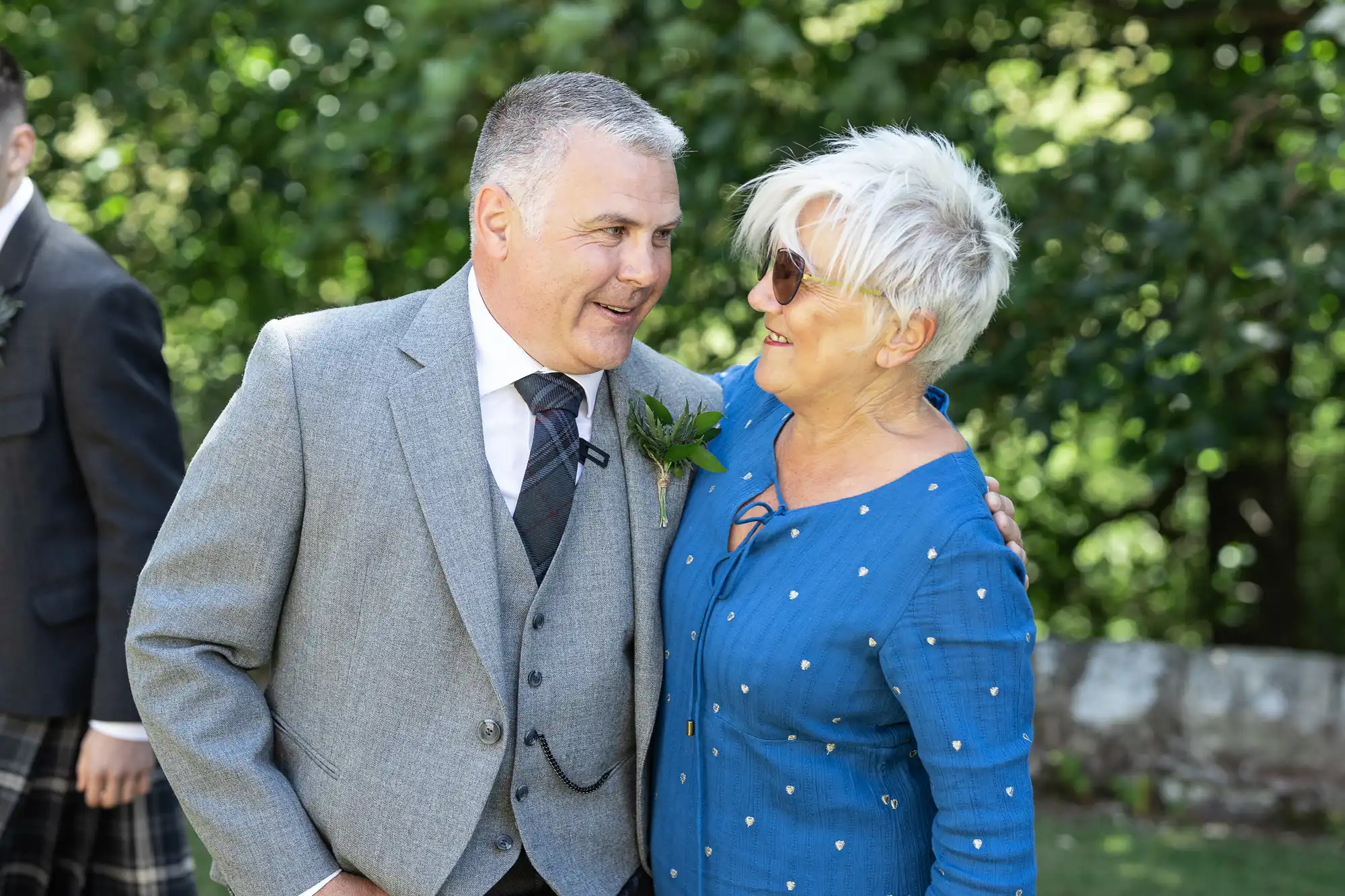 A man in a grey suit and tie smiles while standing next to a woman in a blue dress and sunglasses. They are outdoors with greenery in the background.