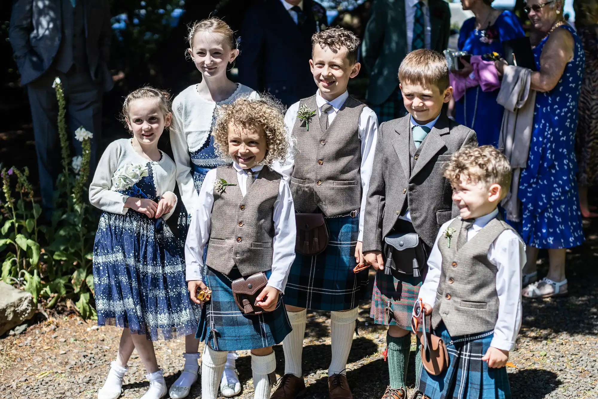 Seven children, dressed in formal attire with some wearing kilts, pose together for a group photo outdoors with other people in the background.