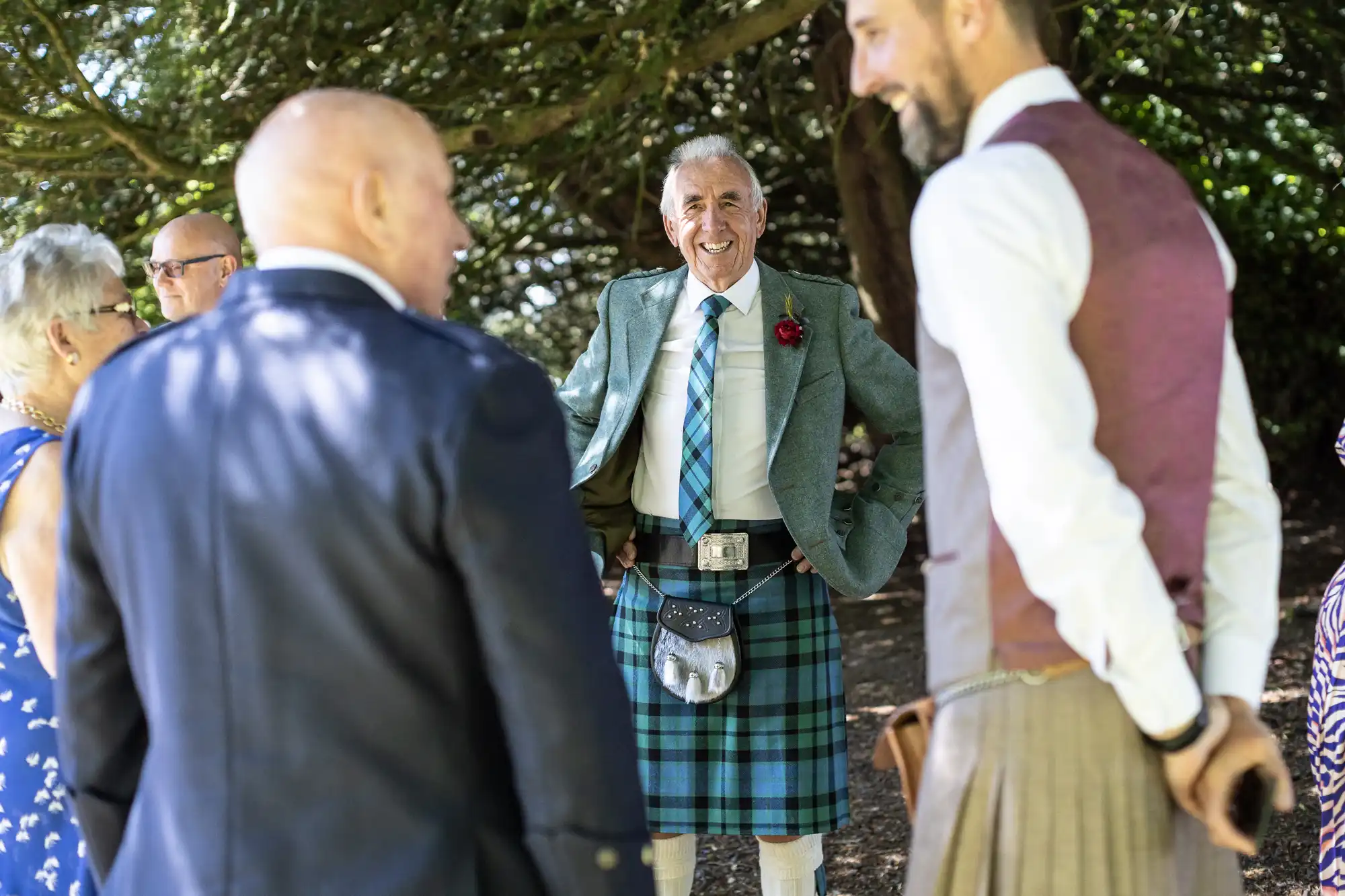 An older man wearing a green jacket and plaid kilt smiles and mingles with other formally dressed individuals in an outdoor setting.