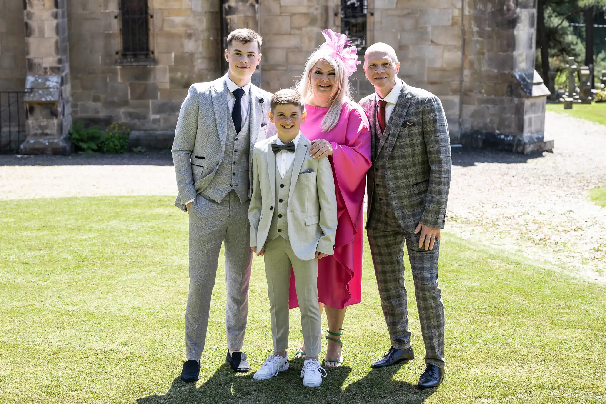 A family of four, dressed in formal attire, stands together on a lawn in front of a stone building. Two boys wear light-colored suits, the woman wears a bright pink dress with a fascinator, and the man wears a plaid suit.