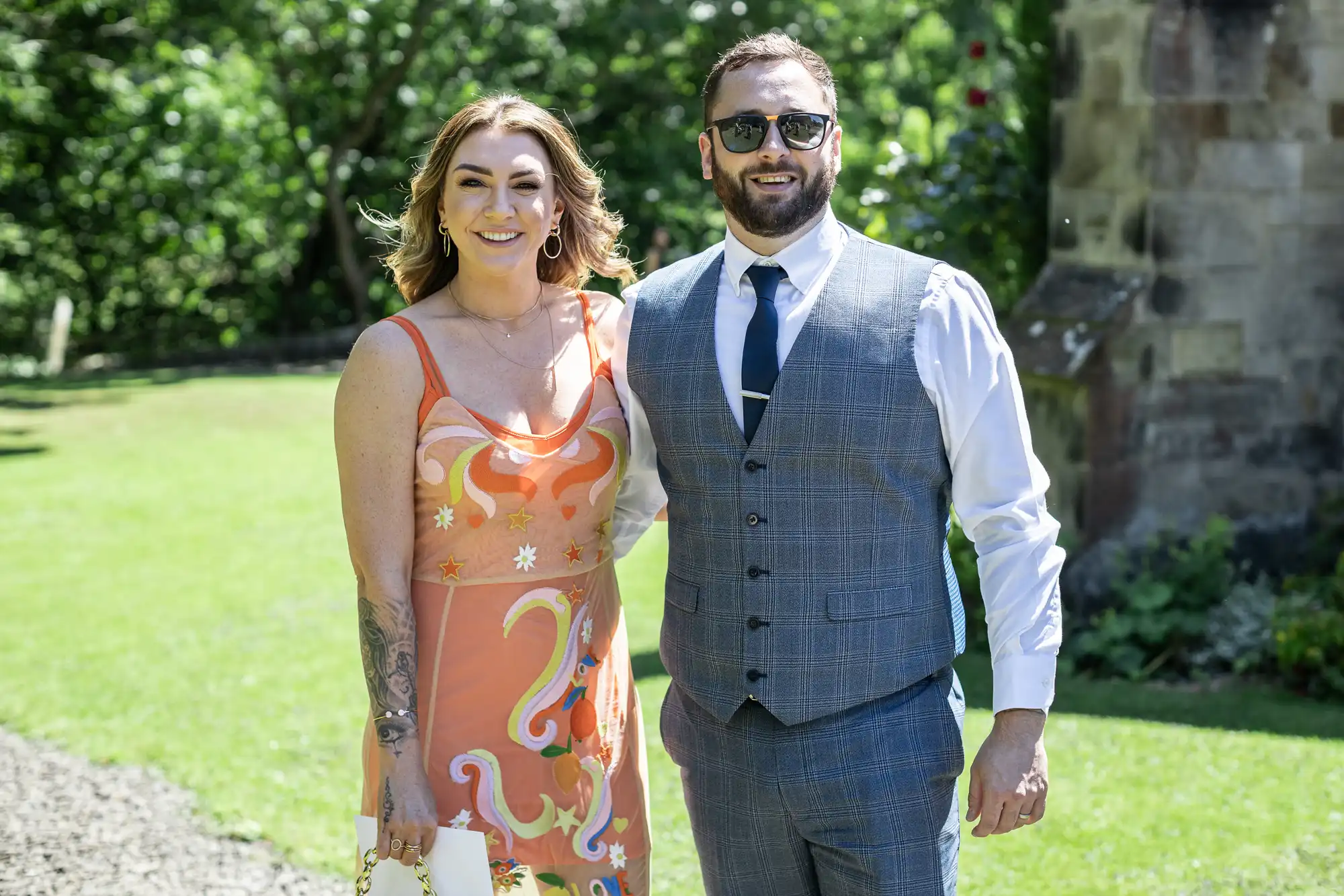 A woman in an orange floral dress and a man in a grey suit with sunglasses stand together outside on a sunny day.