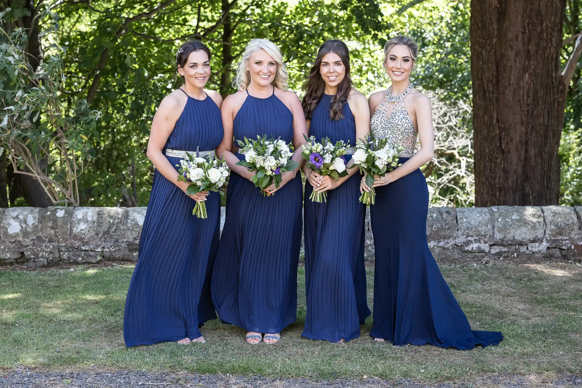 Four women in navy blue dresses stand outdoors, holding white and green floral bouquets, posing for a photo against a backdrop of trees and stone wall.