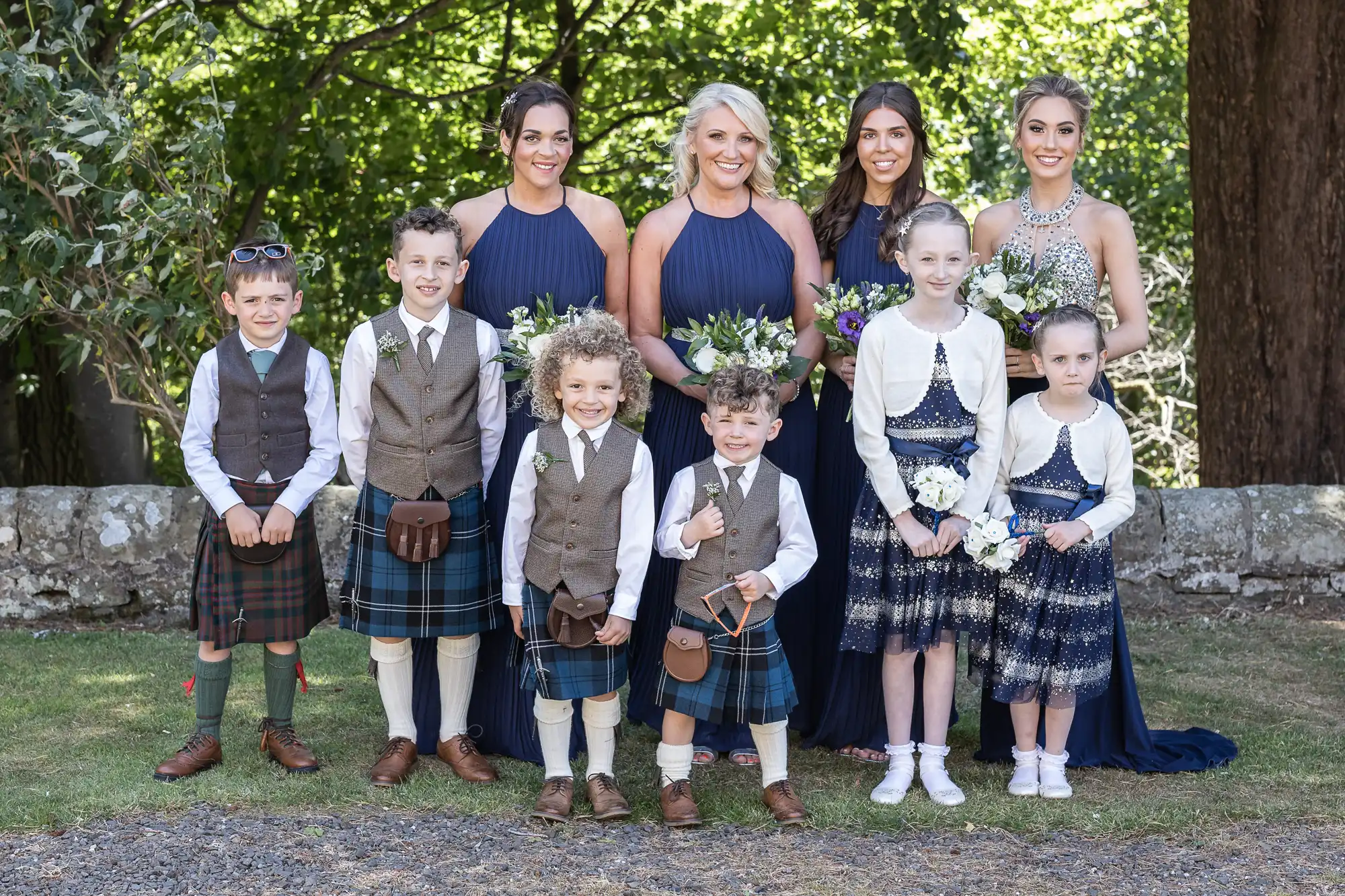 A group photo of four young boys, four young girls, and four adult women, all dressed in formal attire, posed outdoors in front of a stone wall with greenery in the background.