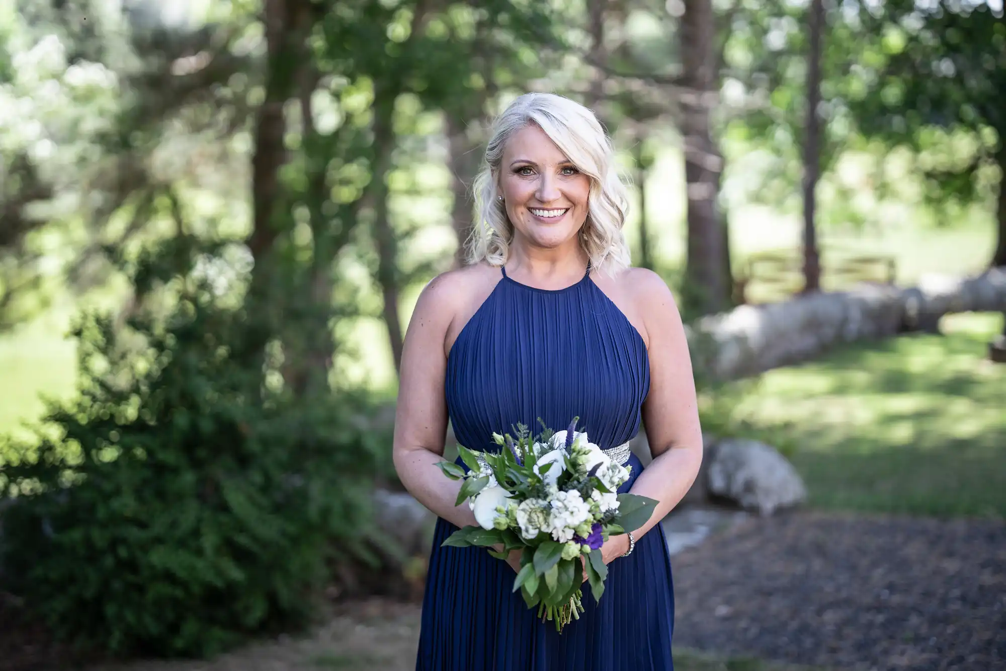 A woman in a navy blue dress holding a bouquet of white and purple flowers stands outdoors with trees in the background.