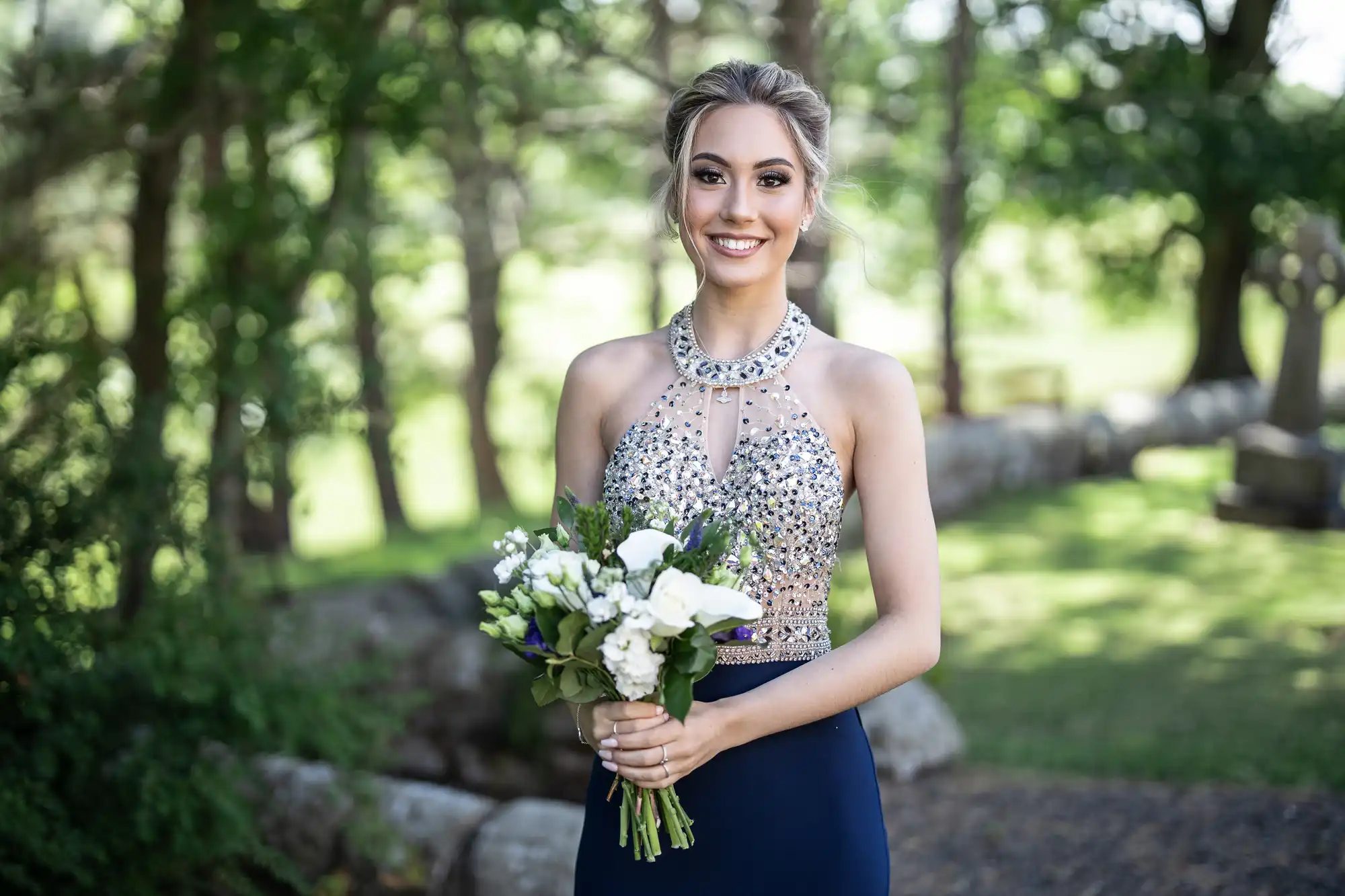 A person in a sparkly dress holds a bouquet of flowers while standing outdoors with trees in the background.