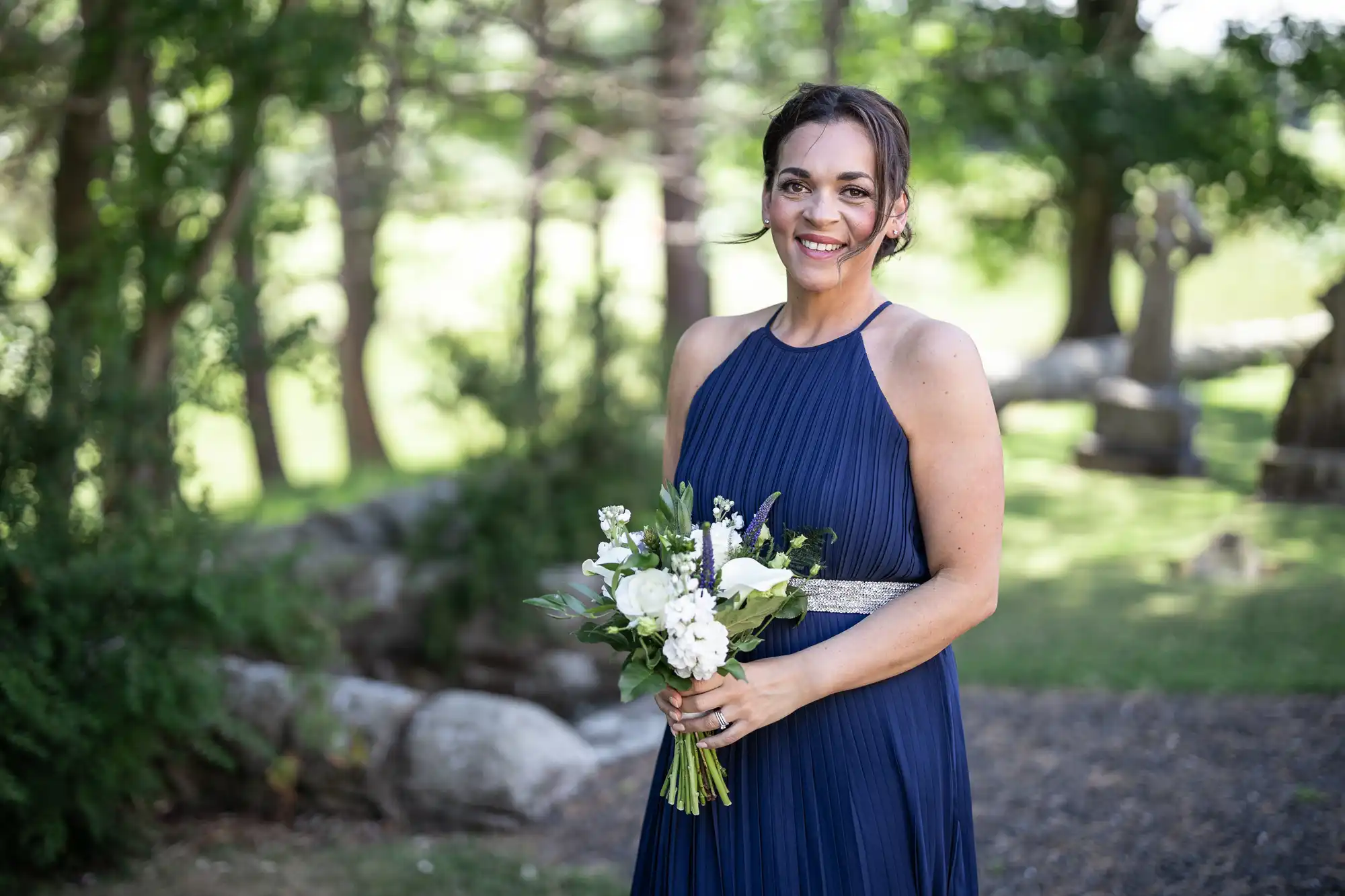 A woman in a sleeveless navy blue dress holds a bouquet of white and green flowers while standing outdoors in a park.