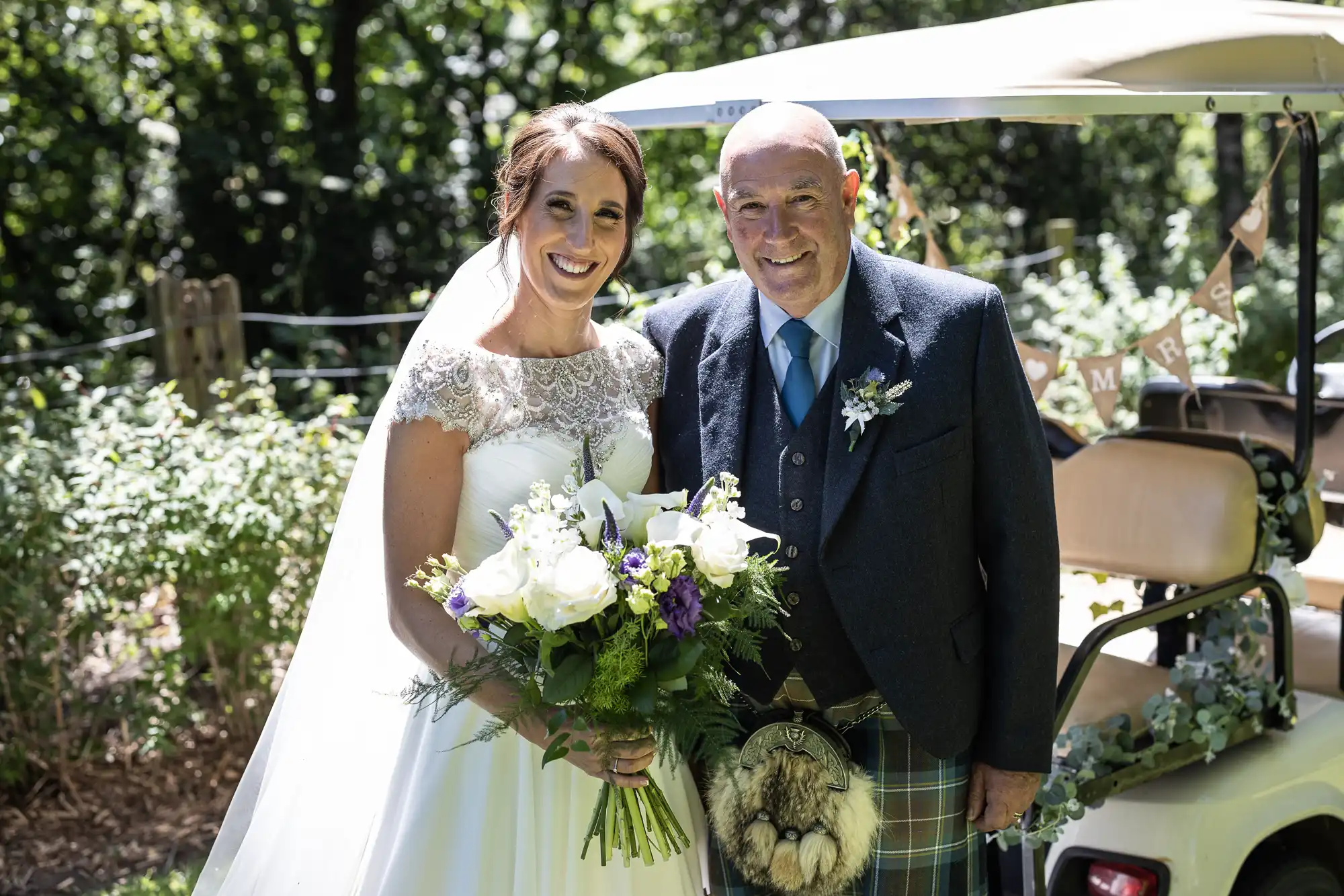 A bride in a white dress and veil stands next to an older man in a suit and kilt, holding a bouquet of flowers. They are outdoors in a garden setting with a decorated golf cart in the background.