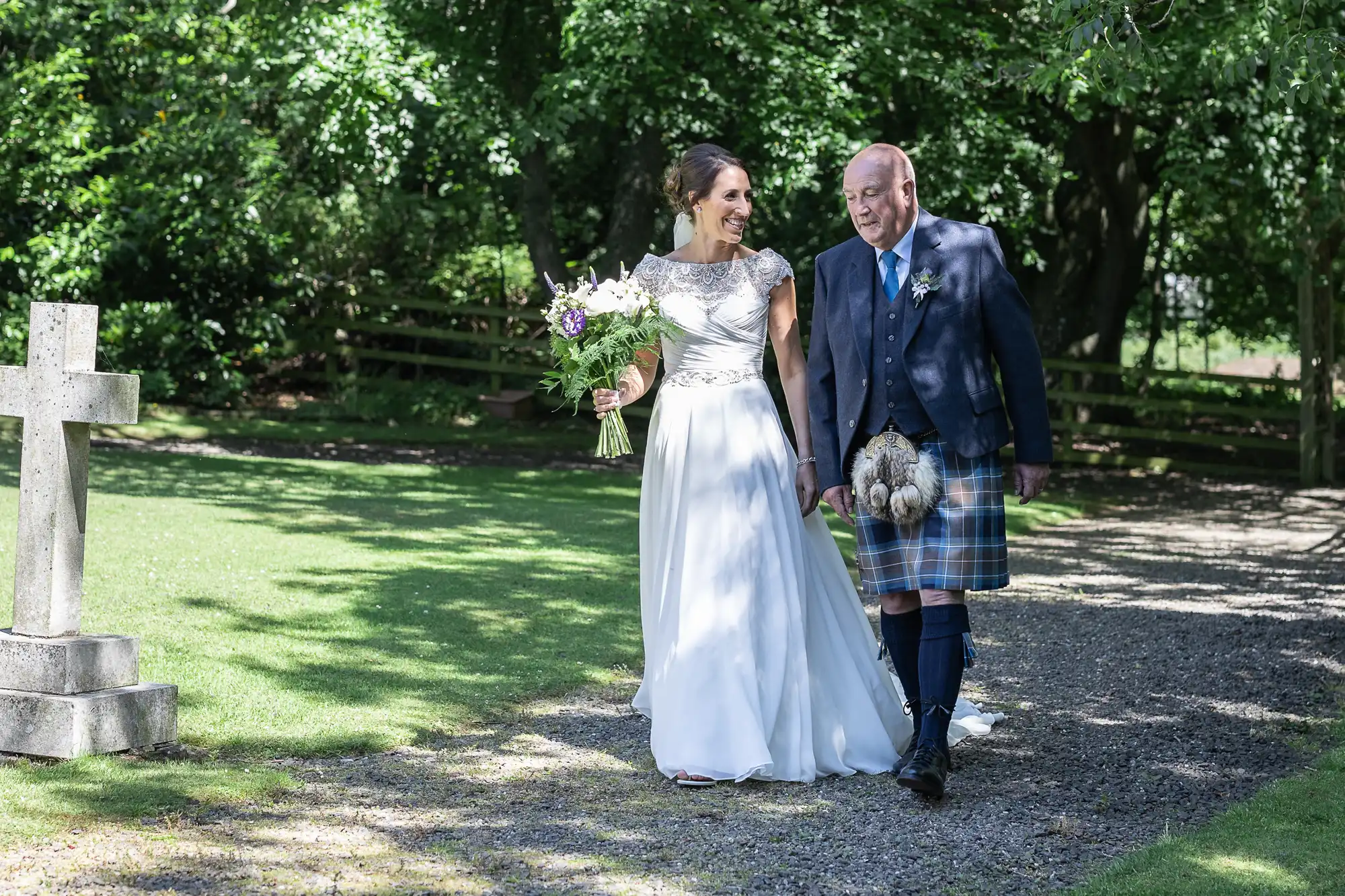 A bride in a white dress and a man in a kilt walk hand in hand outdoors, surrounded by greenery and sunlight.