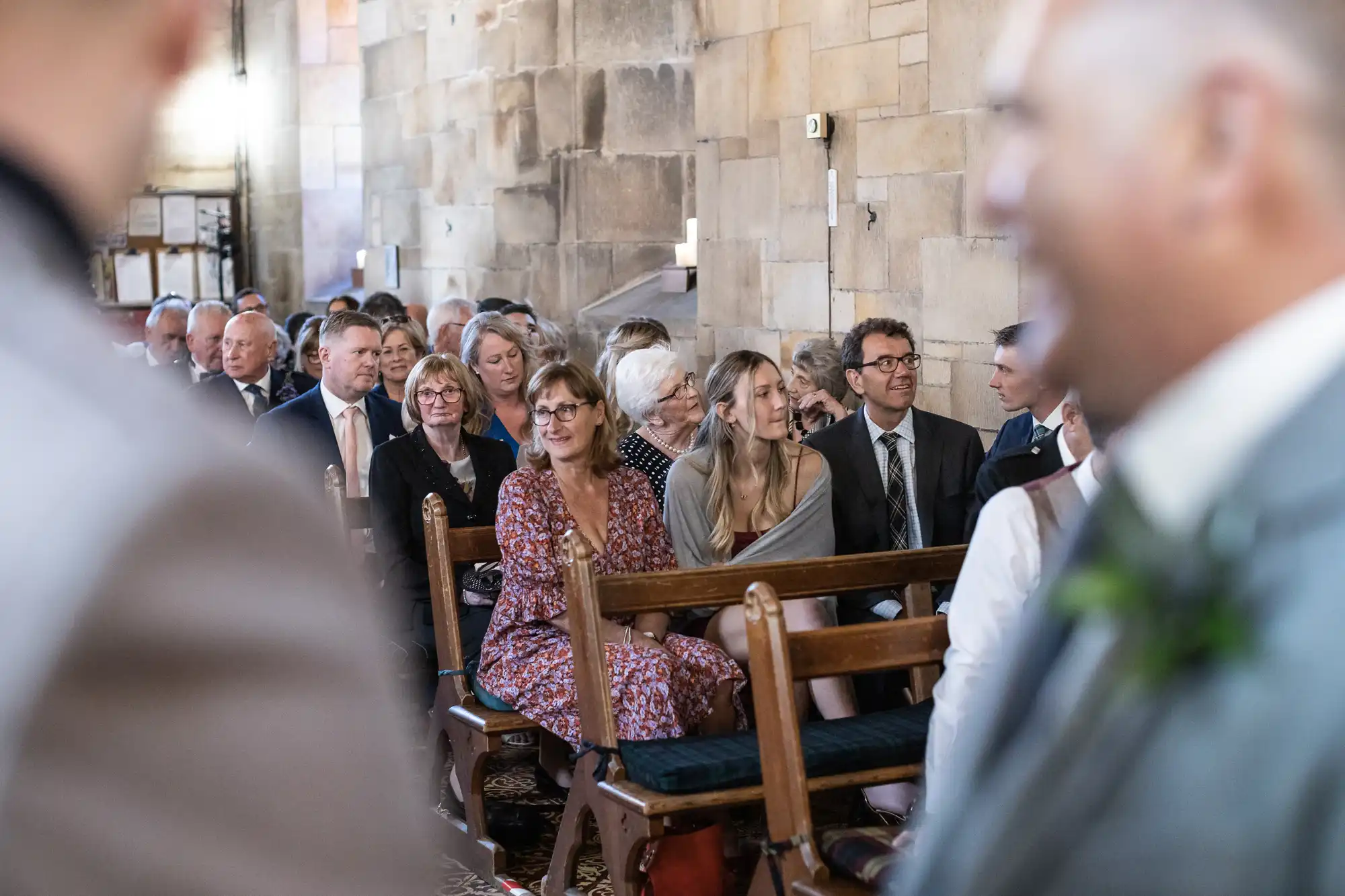 A group of people sit on wooden benches inside a stone-walled building, attentively facing towards the front where an event appears to be occurring.