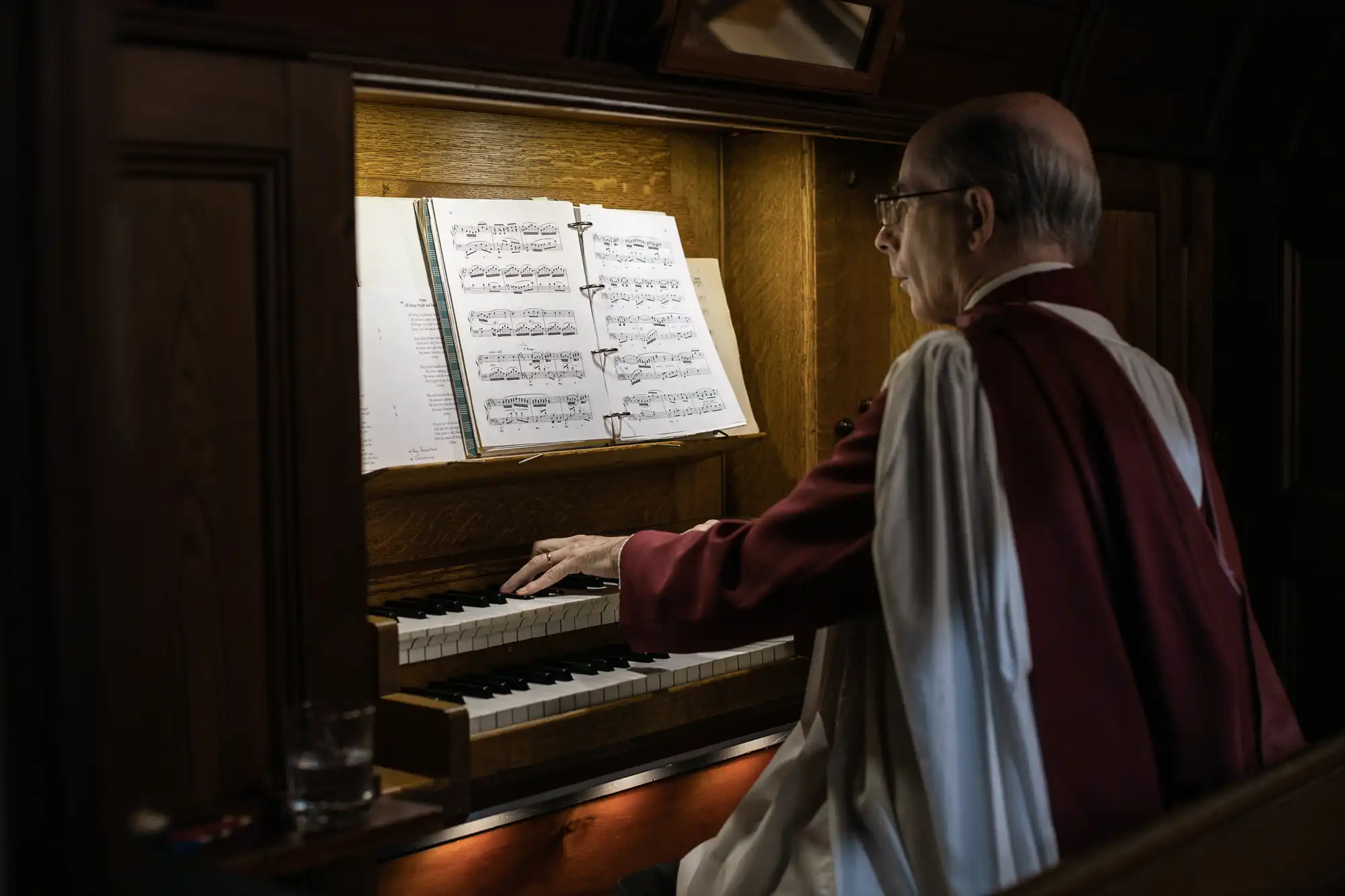 A person in a red and white robe plays an organ, reading sheet music propped up on the instrument.