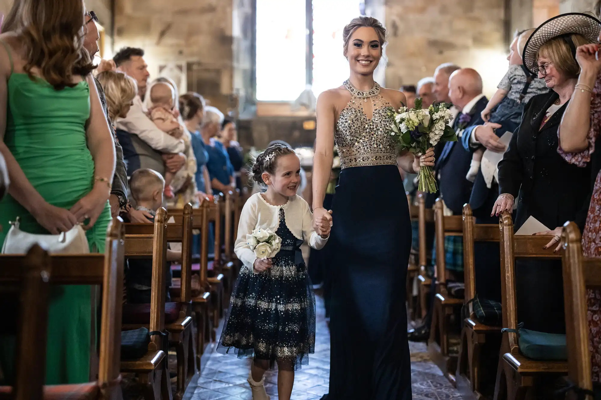 A woman in a formal gown and a young girl walk down the aisle of a church, surrounded by seated and standing guests. The woman holds a bouquet, and the girl carries a small arrangement of flowers.