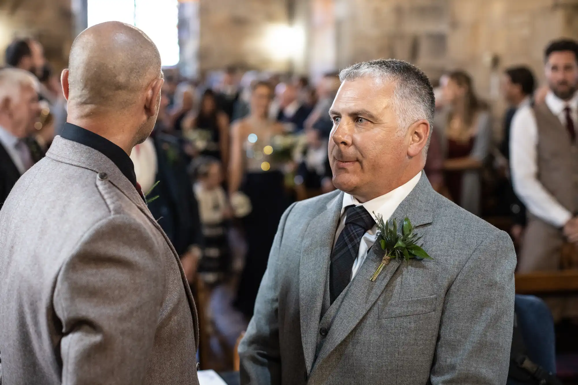 Two men in formal attire stand face to face in a church, surrounded by other people. One man has gray hair and a boutonniere, while the other is bald.