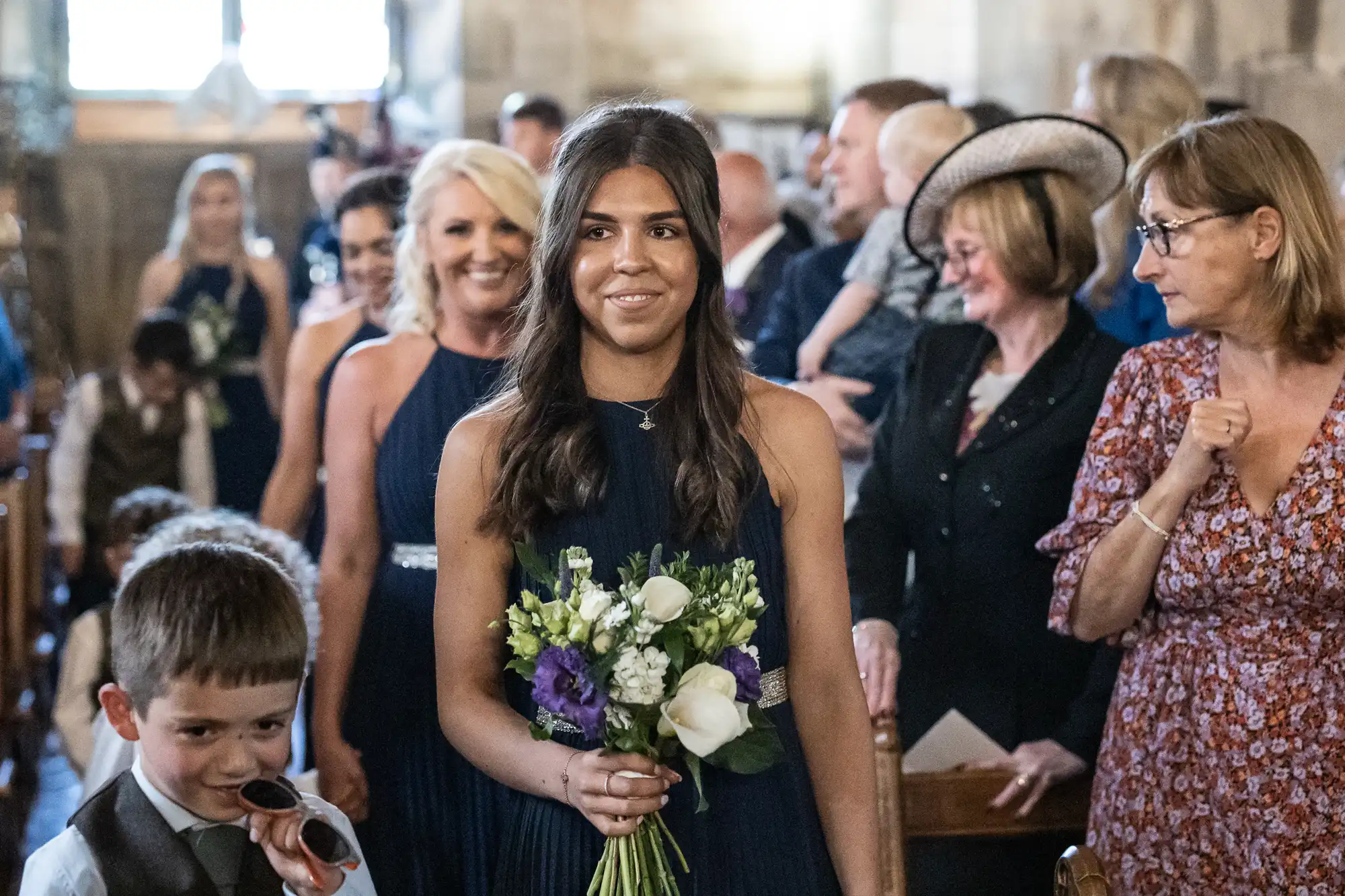 A young woman holding a bouquet walks down the aisle in a church, followed by other people. She is wearing a dark dress and has long brown hair. People in the audience watch and smile.