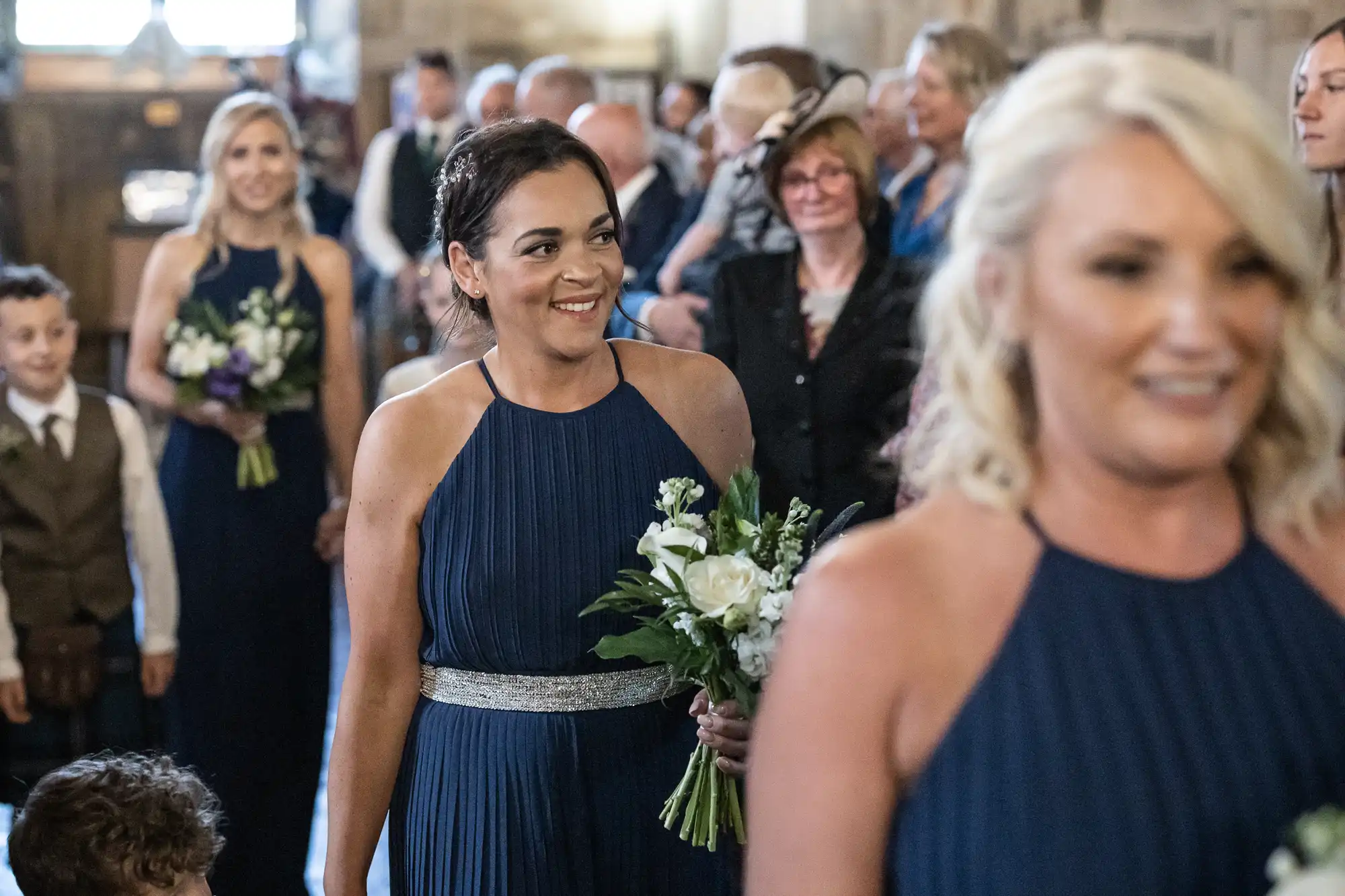 Women in navy blue dresses, holding bouquets, walk down an aisle at an indoor event; the seated crowd watches.