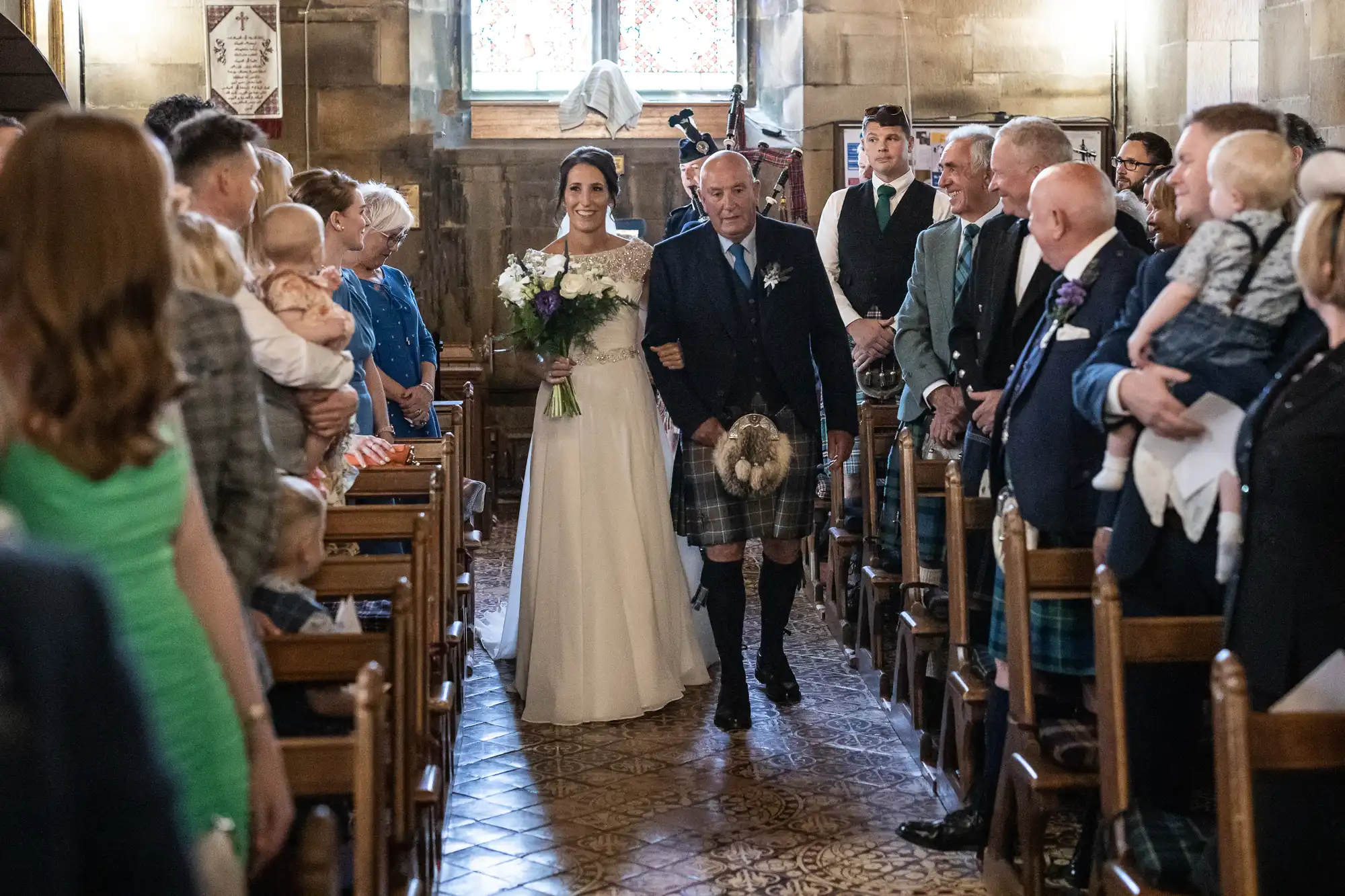 A bride holding a bouquet walks down the aisle in a church, accompanied by an older man in a kilt. Guests seated on wooden benches look on.