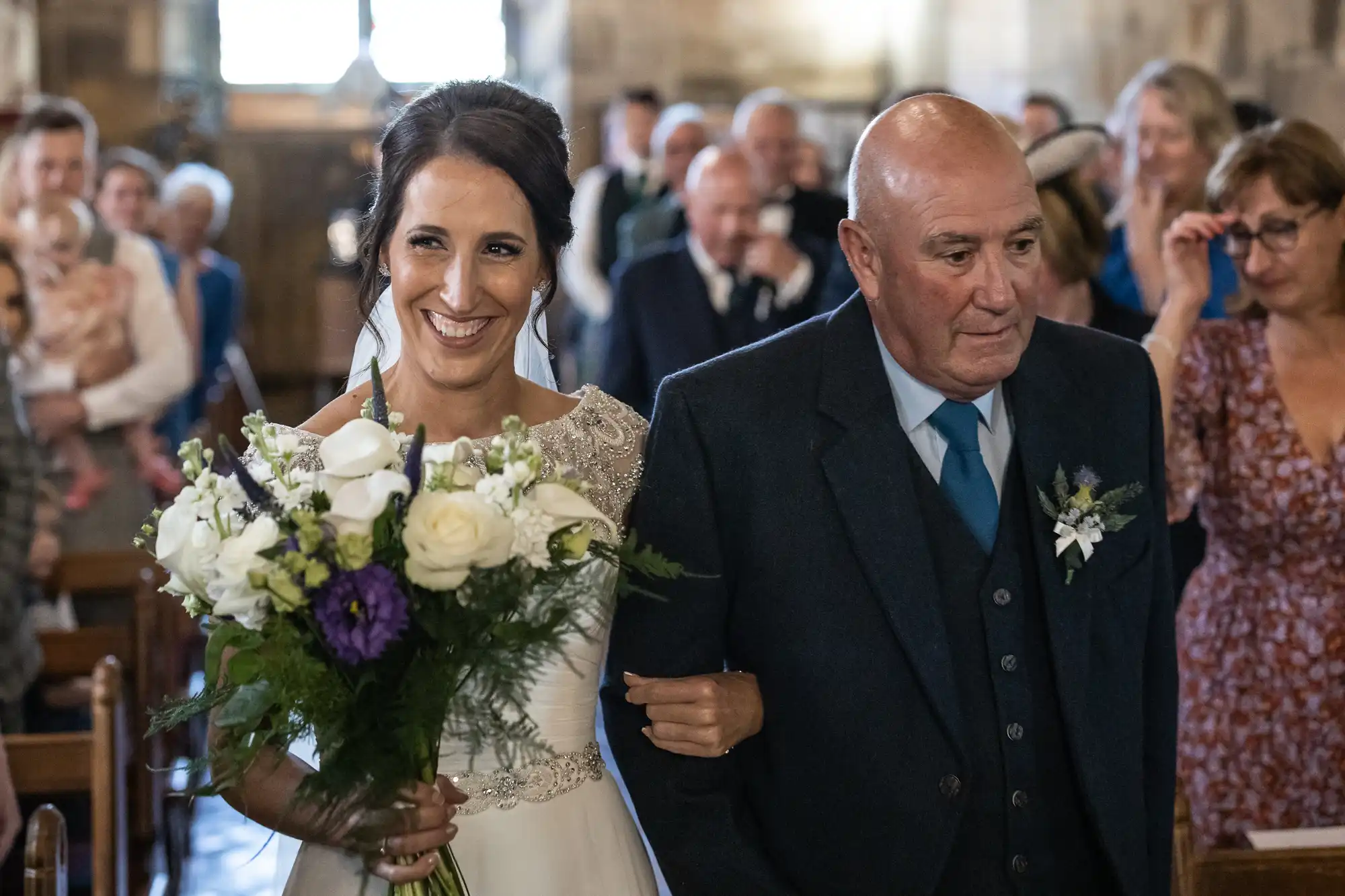 A bride holding a floral bouquet walks down the aisle with an older man at a wedding ceremony, with guests in the background.