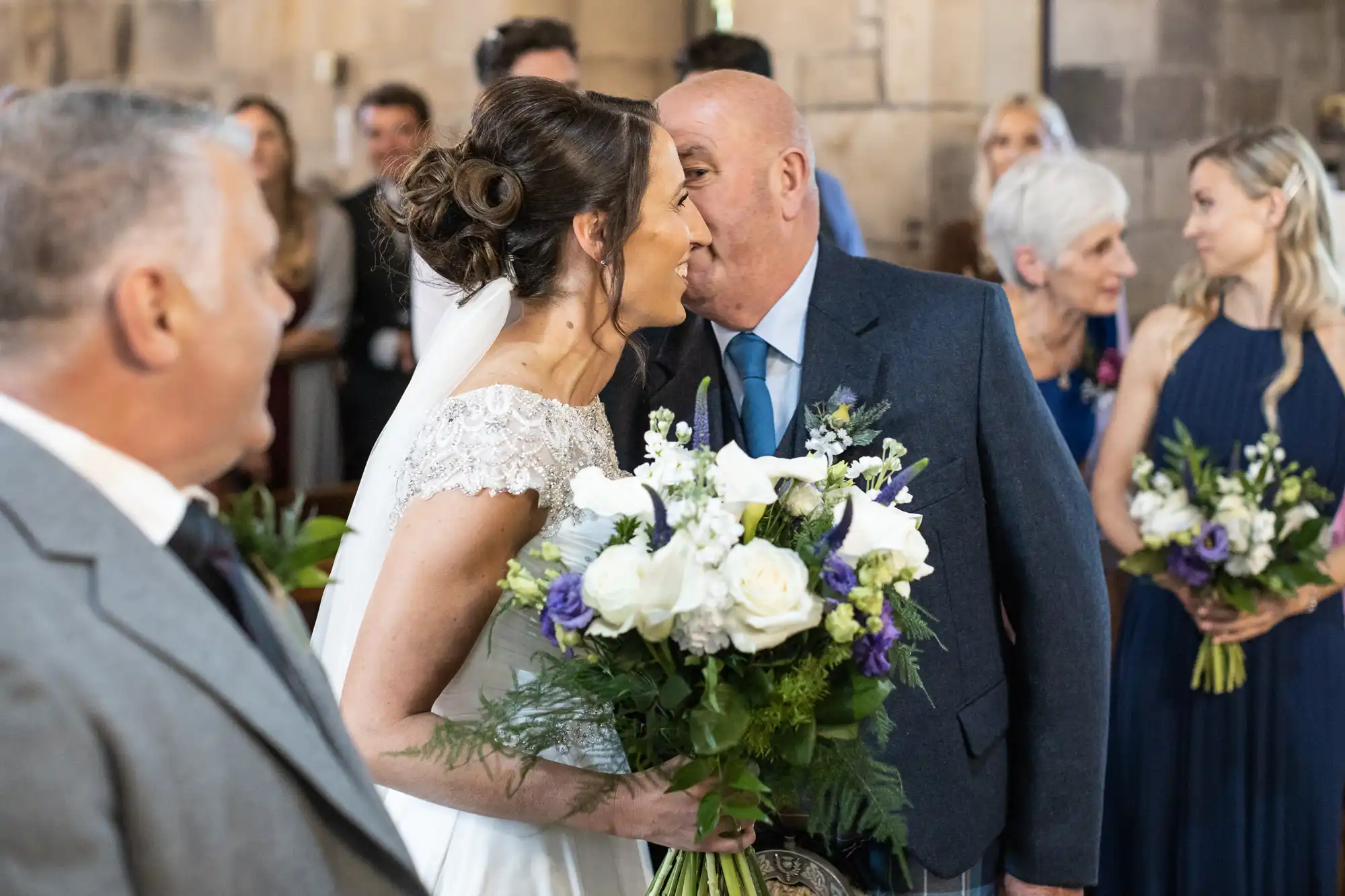 A bride holding a bouquet shares a kiss with an older man, possibly her father, inside a stone-walled venue. Other people in formal attire are present in the background.