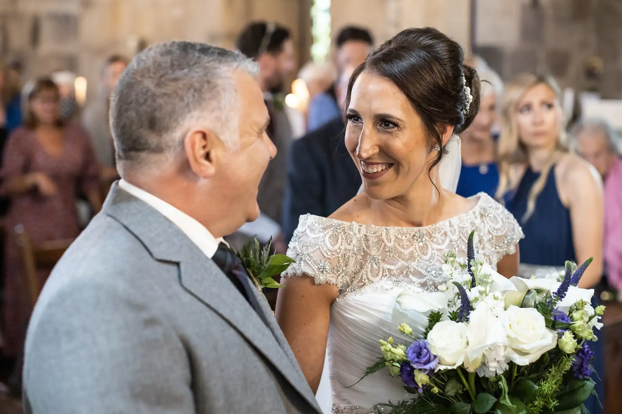 A bride and groom smile at each other during their wedding ceremony, with guests in the background. The bride holds a floral bouquet.