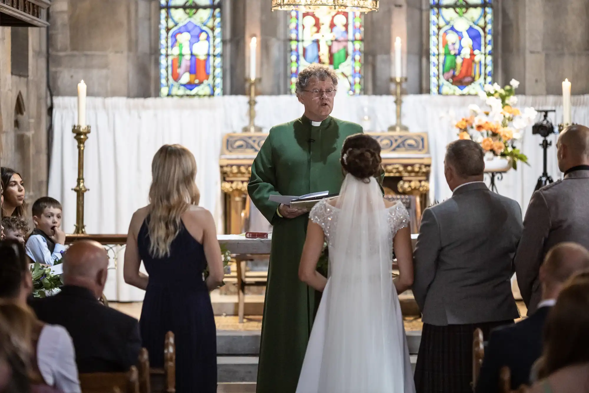 A wedding ceremony is taking place in a church with a priest in a green robe standing behind the altar, and a bride and groom facing him. Stained glass windows are visible in the background.