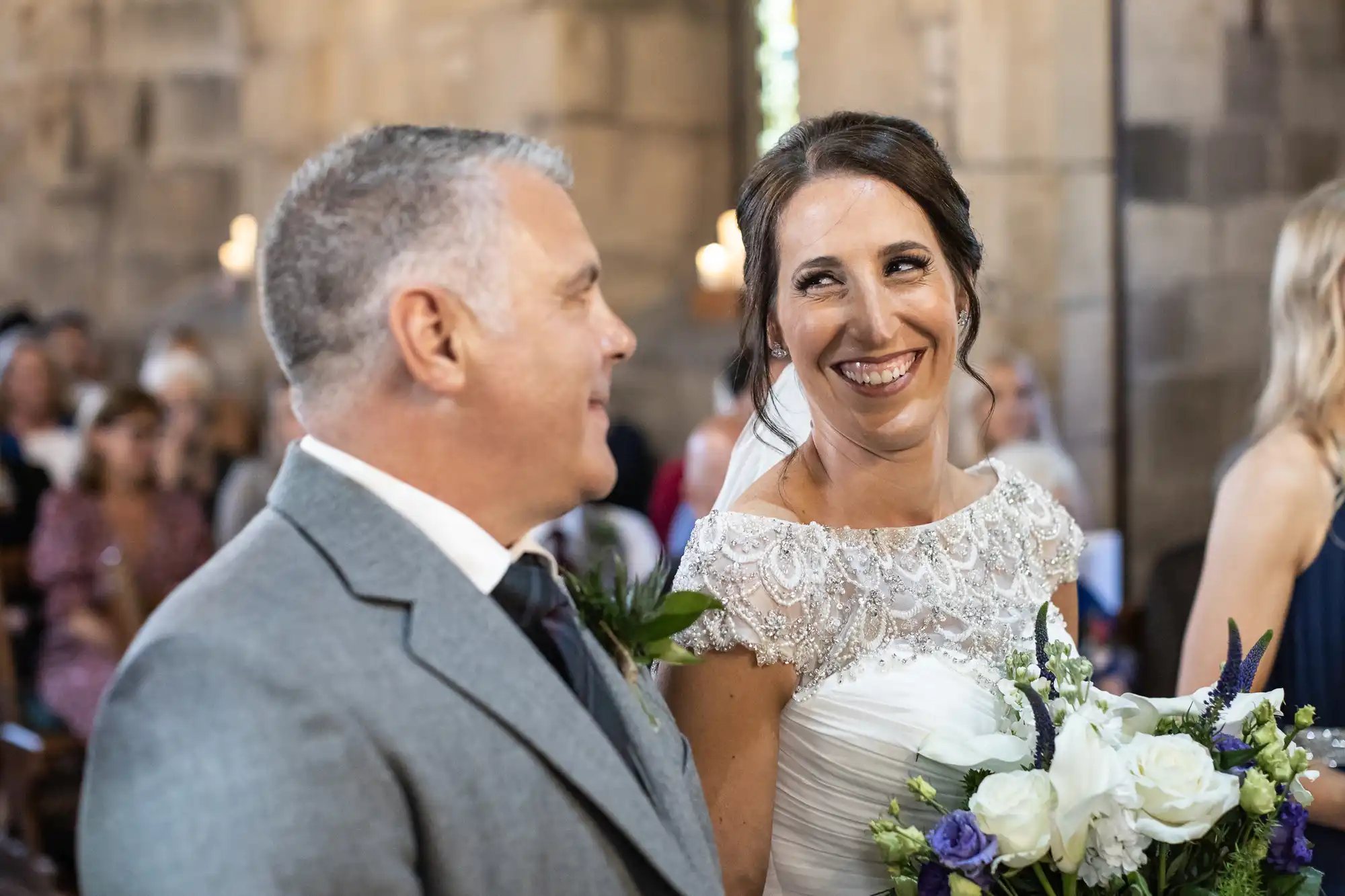 The bride smiles at a man in a suit while holding a bouquet of flowers. They are inside a stone building with people seated in the background.