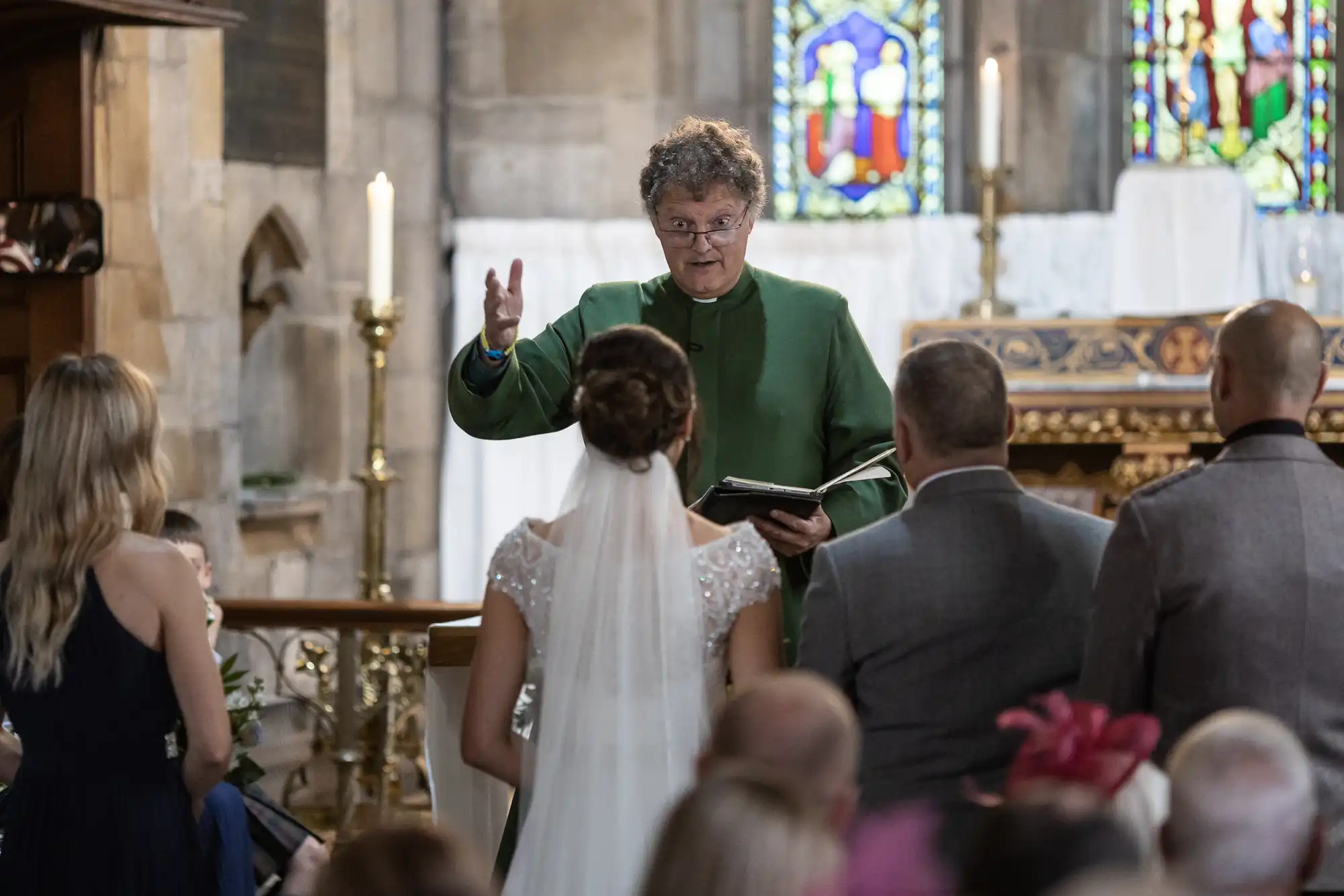 A person in a green robe addresses a wedding couple in a church. Guests are seated, and stained glass windows and candles are visible in the background.