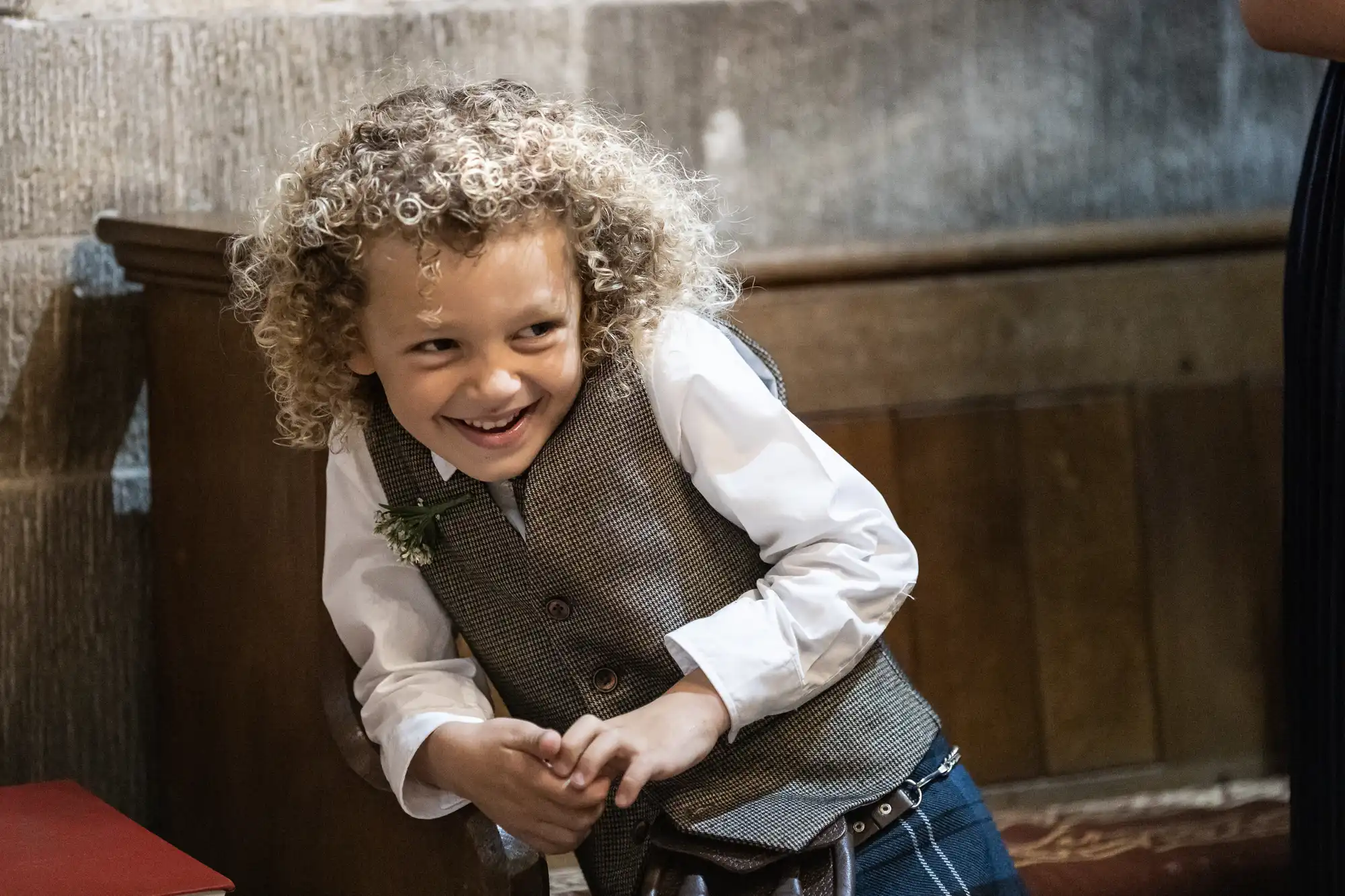 A young child with curly hair, wearing a white shirt and grey vest, leans on a wooden bench indoors, smiling and looking playfully to the side.