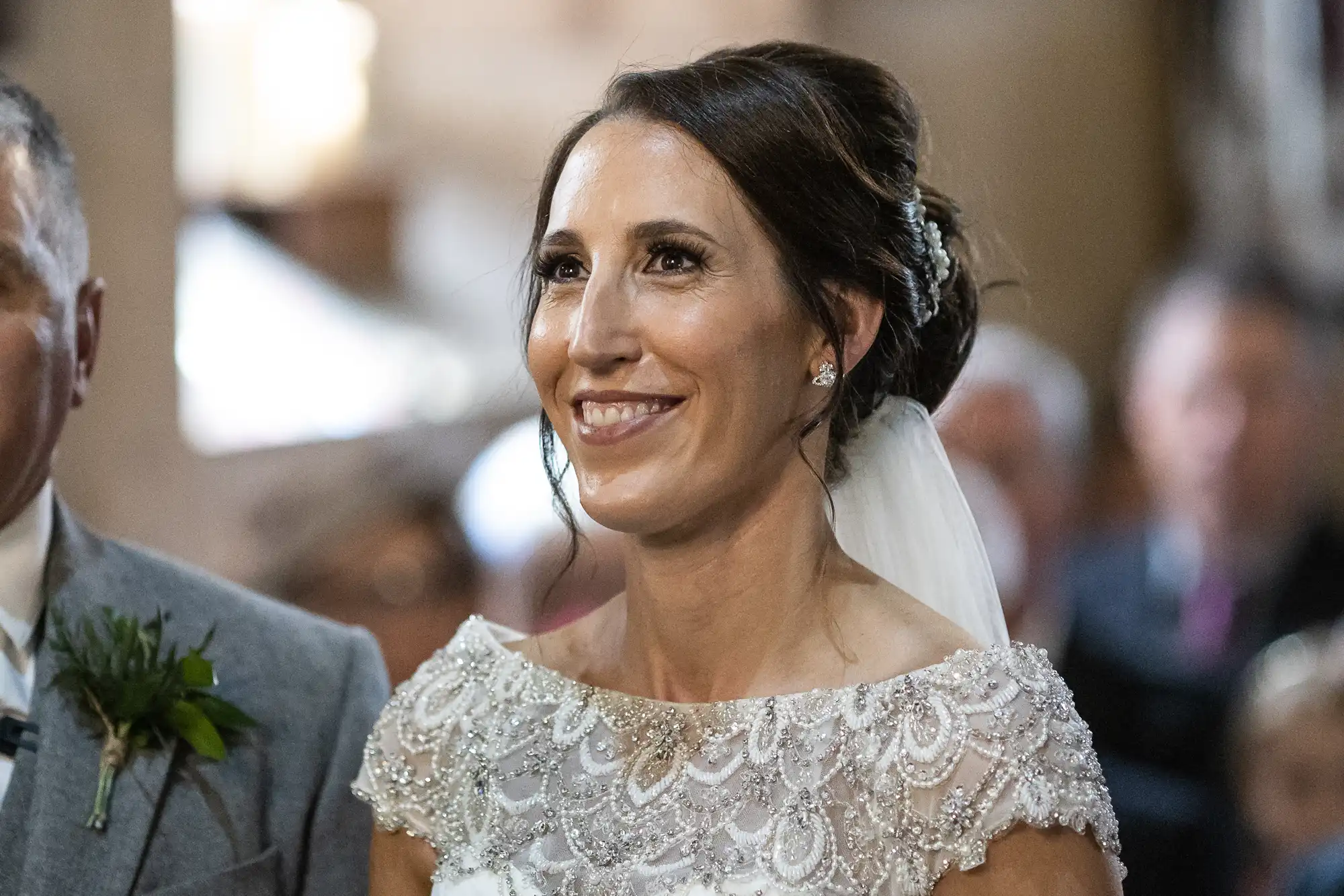 A bride wearing a lace wedding dress and veil smiles as she stands next to a person in a suit, with a blurred background of wedding guests.