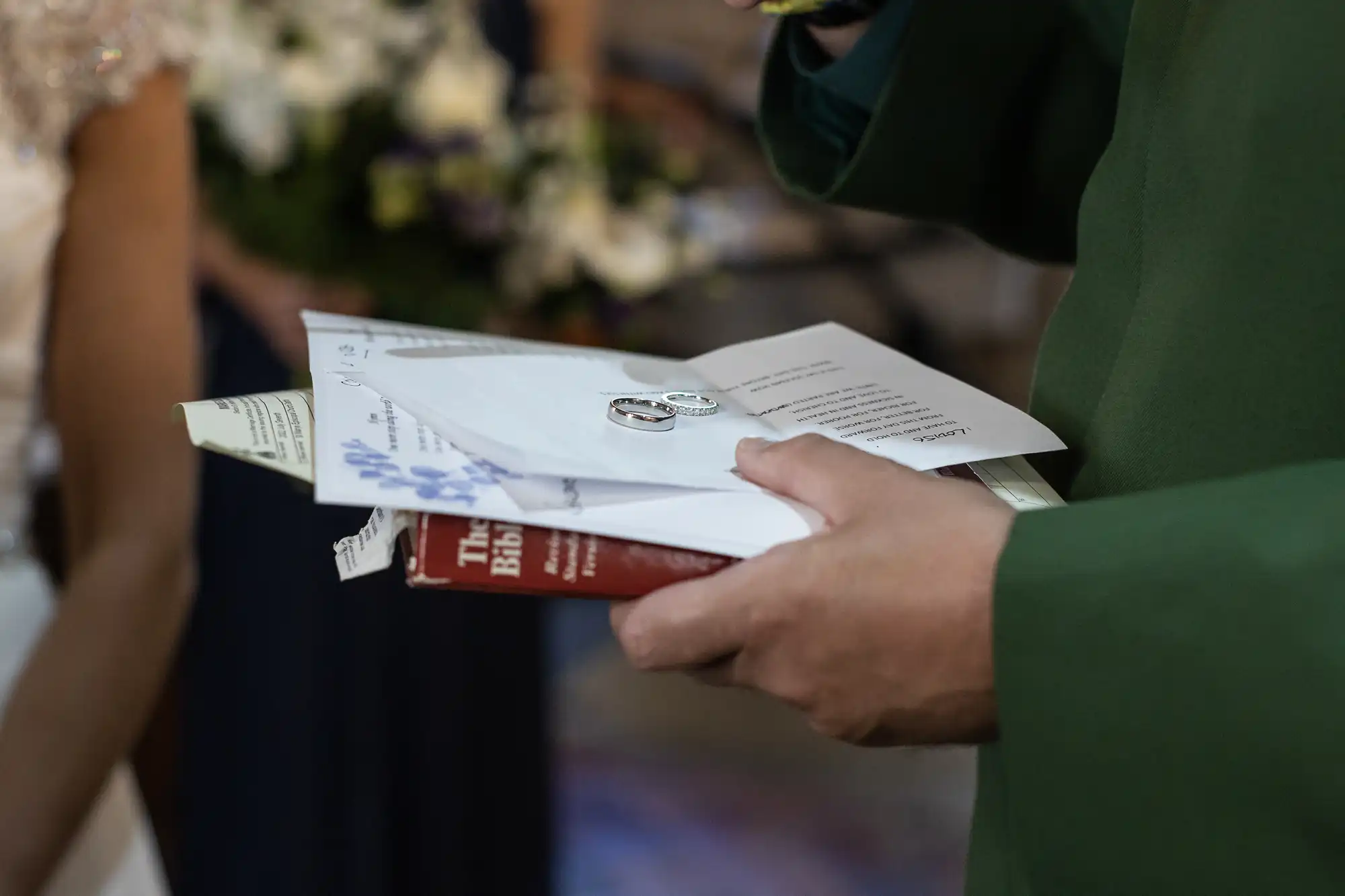 A person holds a small stack of items including a booklet, documents, two rings, and a red book titled "The Bible" during a ceremony.