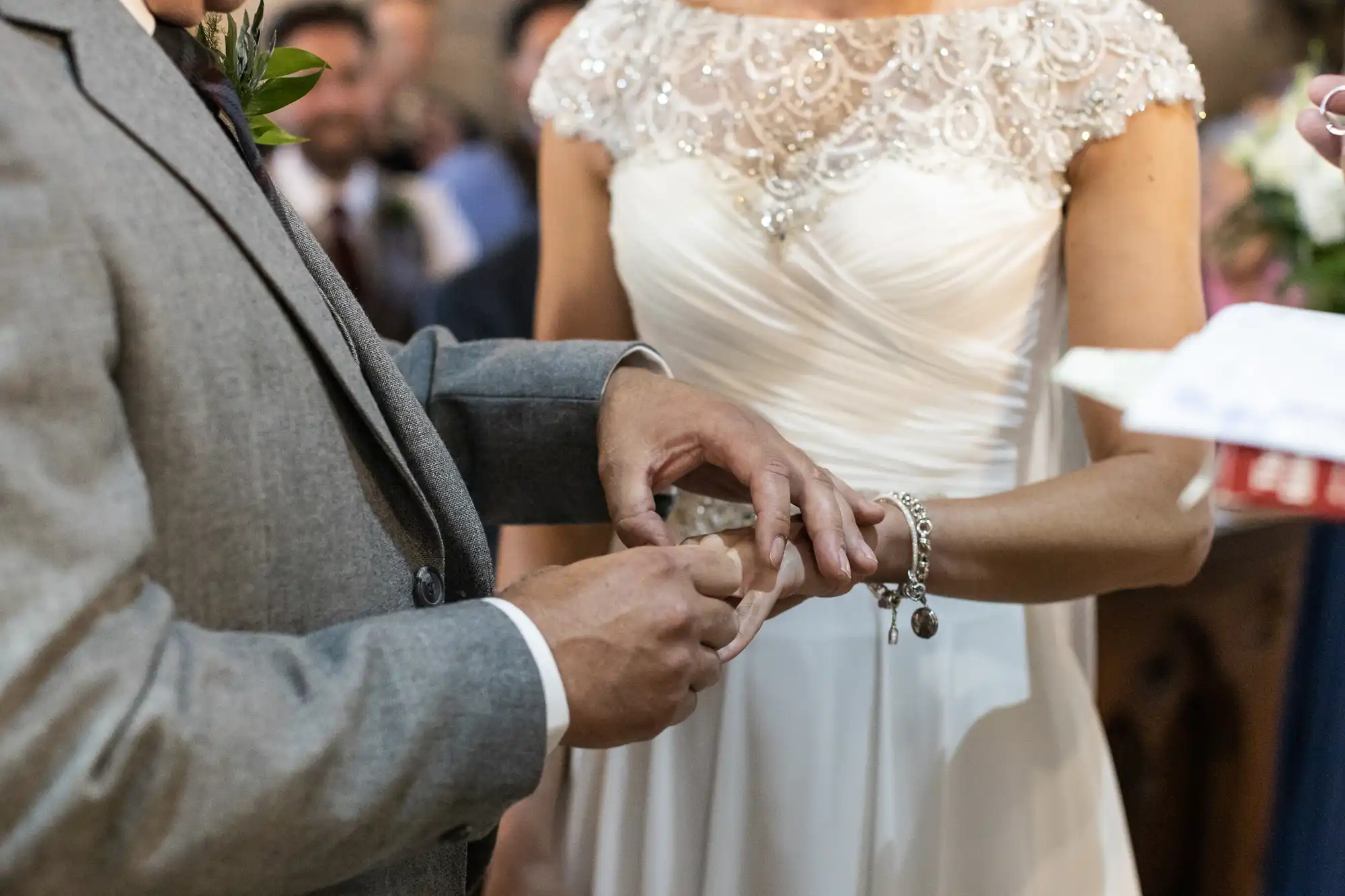 A groom places a ring on the bride's finger during a wedding ceremony. Both are dressed in formal attire; the bride is wearing a white dress and the groom a gray suit.