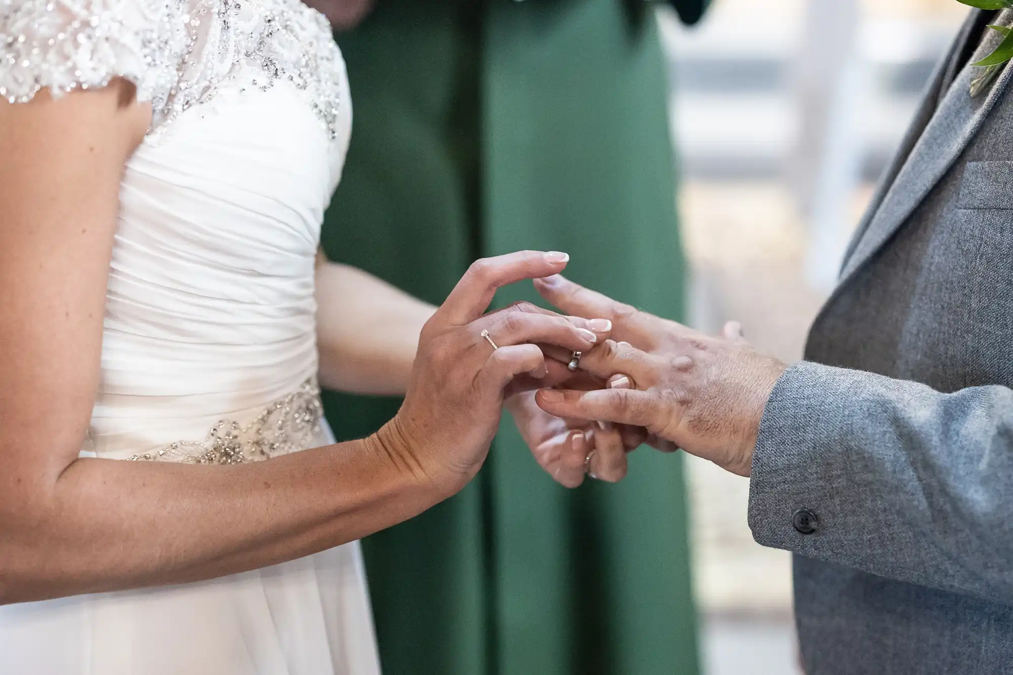 A close-up of a bride placing a ring on the groom’s finger during a wedding ceremony. The bride is wearing a white dress, and the groom is in a gray suit.