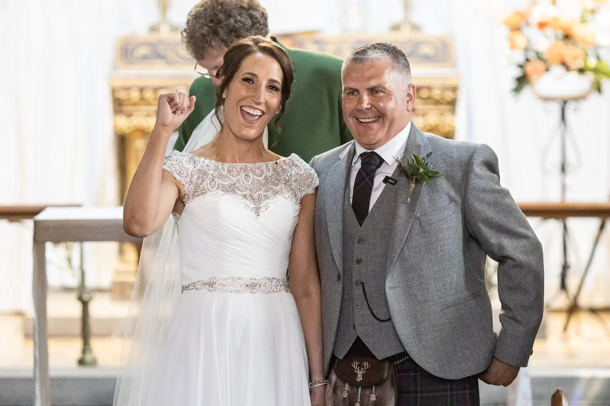 A smiling bride in a white gown and a groom in a grey suit stand together in a church, with the bride raising her hand. A person in green is visible in the background.